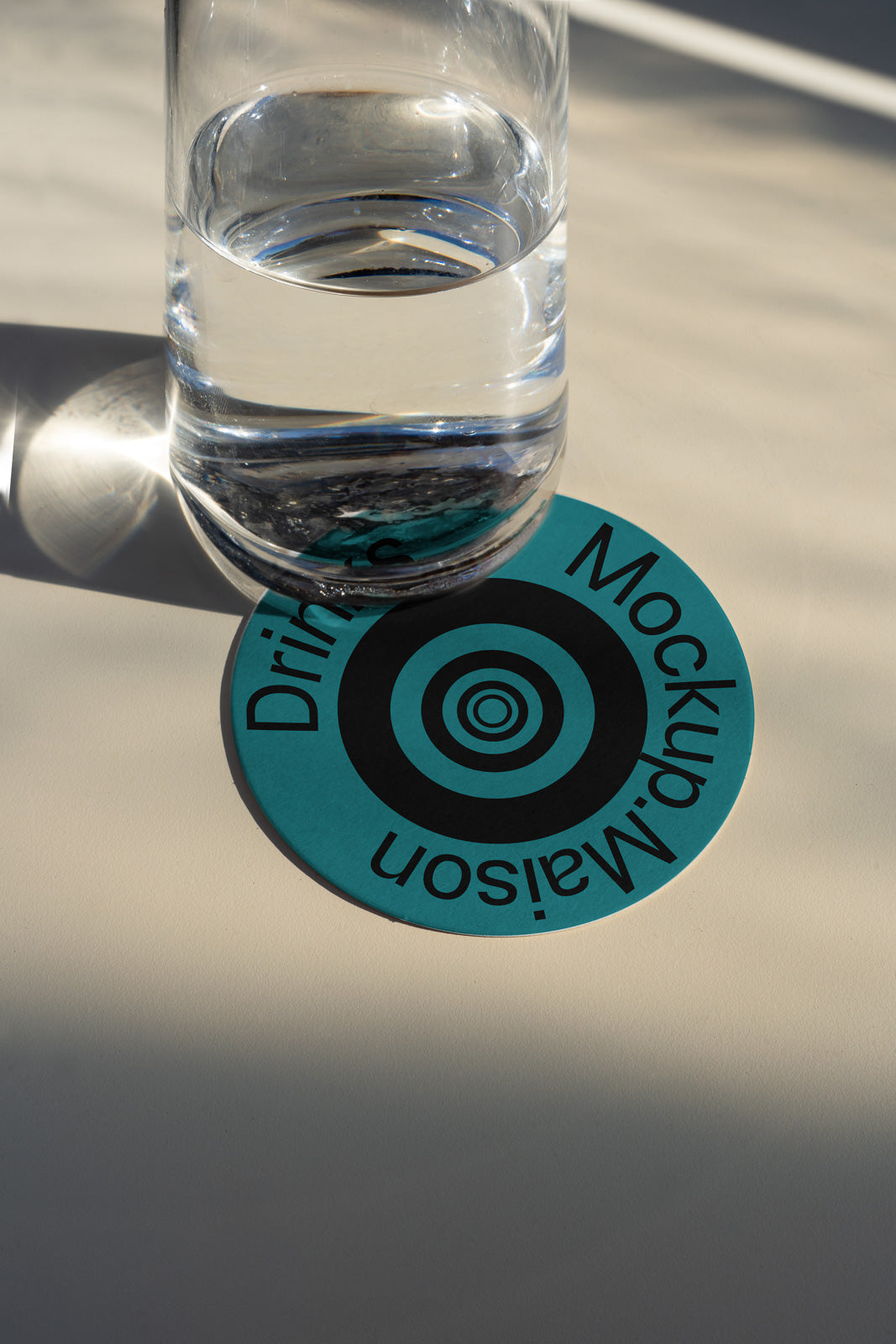 PSD mockup of a clear glass of water placed on a round teal coaster with black concentric circle design and text, casting a shadow on the surface.