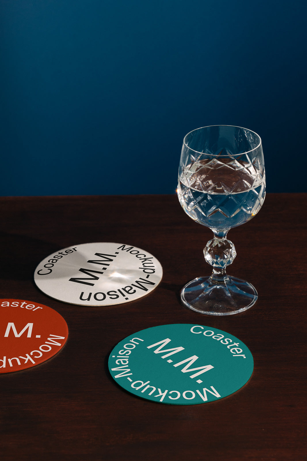 PSD mockup of three branded coasters, each with "M.M. Maison Mockup" text, arranged on a dark wooden surface next to a crystal water glass.