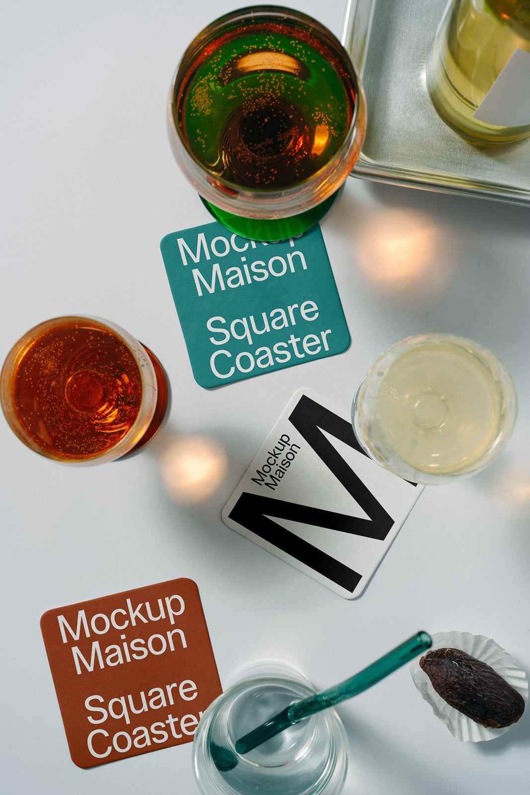 PSD mockup of square coasters under various glasses with drinks, showing "Mockup Maison" branding on a white surface with a date in a wrapper nearby.