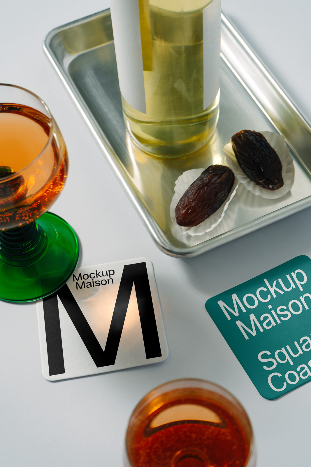PSD mockup of a branding setup featuring a green-stemmed glass with orange liquid, a coaster labeled "Mockup Maison," and a metal tray with a bottle and dates.