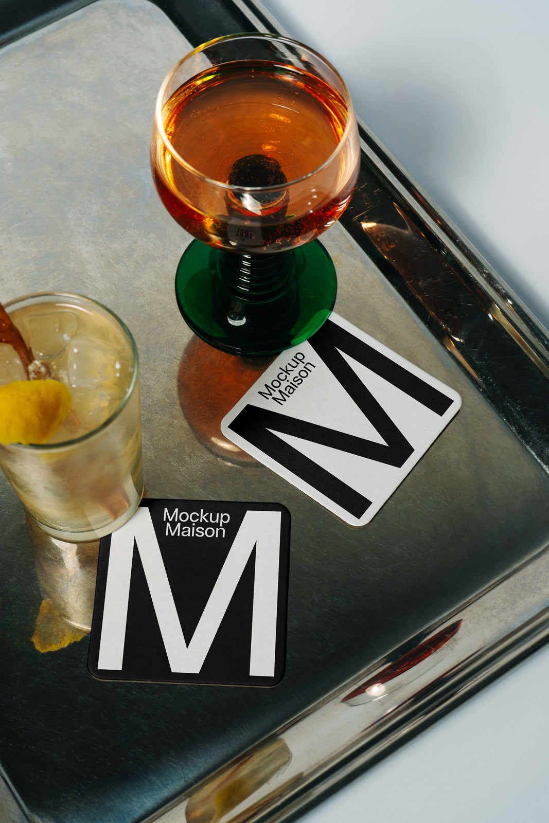 PSD mockup of two coasters with bold letter "M" design placed on a reflective surface next to a glass of whiskey with ice and a cocktail.
