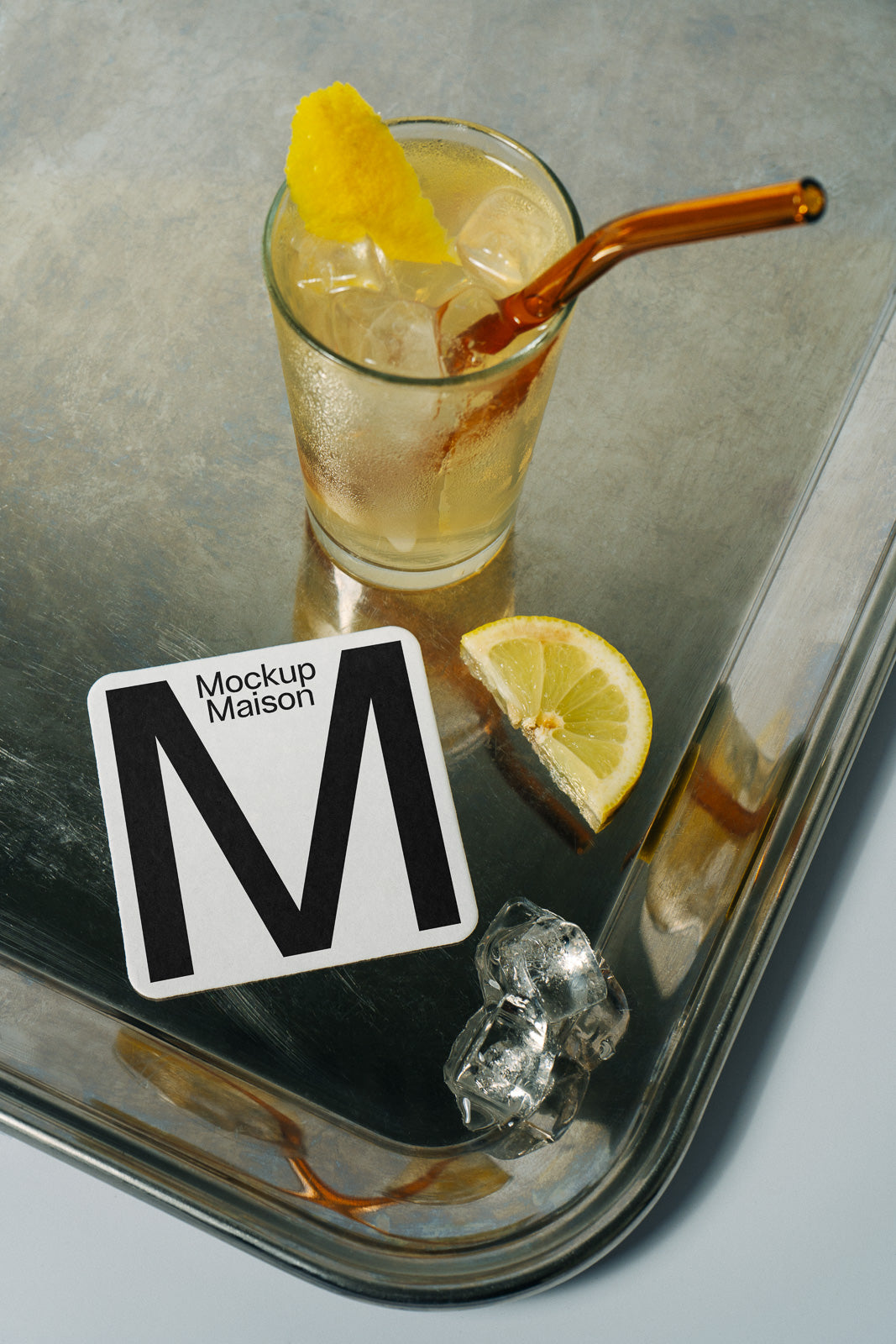 PSD mockup of a cold drink with a glass straw on a metal tray, accompanied by a lemon slice, ice cubes, and a square coaster labeled "Mockup Maison."