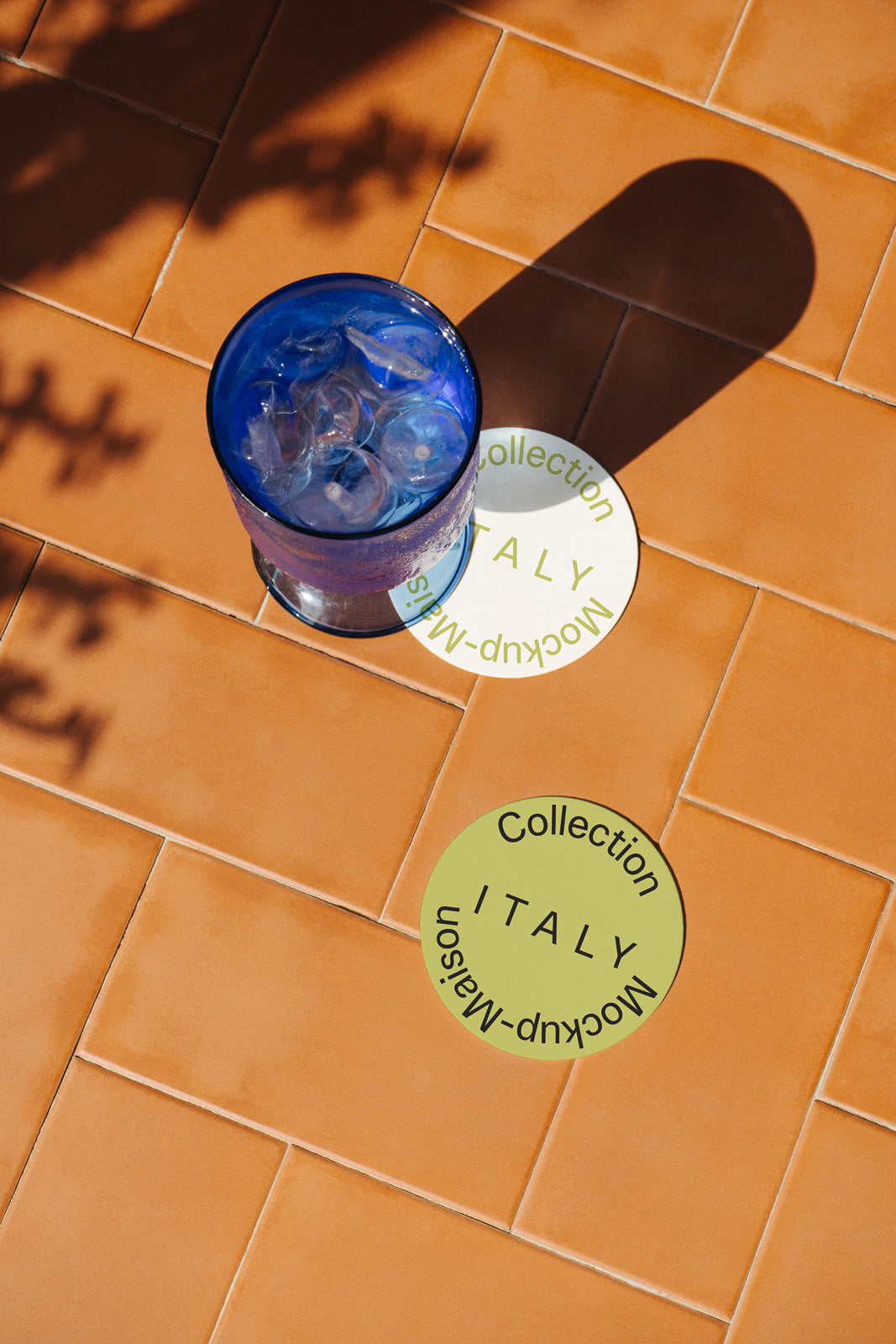 PSD mockup of a blue glass with ice cubes casting a shadow on terracotta tiles, next to round coasters labeled "Collection Italy Mockup Maison."