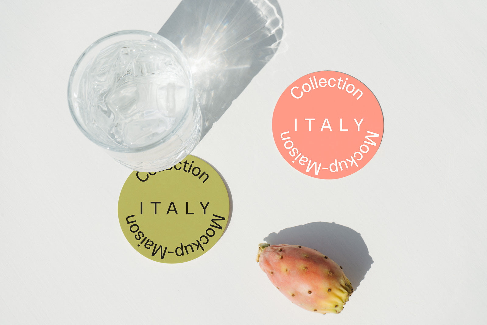 PSD mockup of two circular tags labeled "Collection Italy Mockup-Maison" beside a glass of ice water and a prickly pear on a white surface.