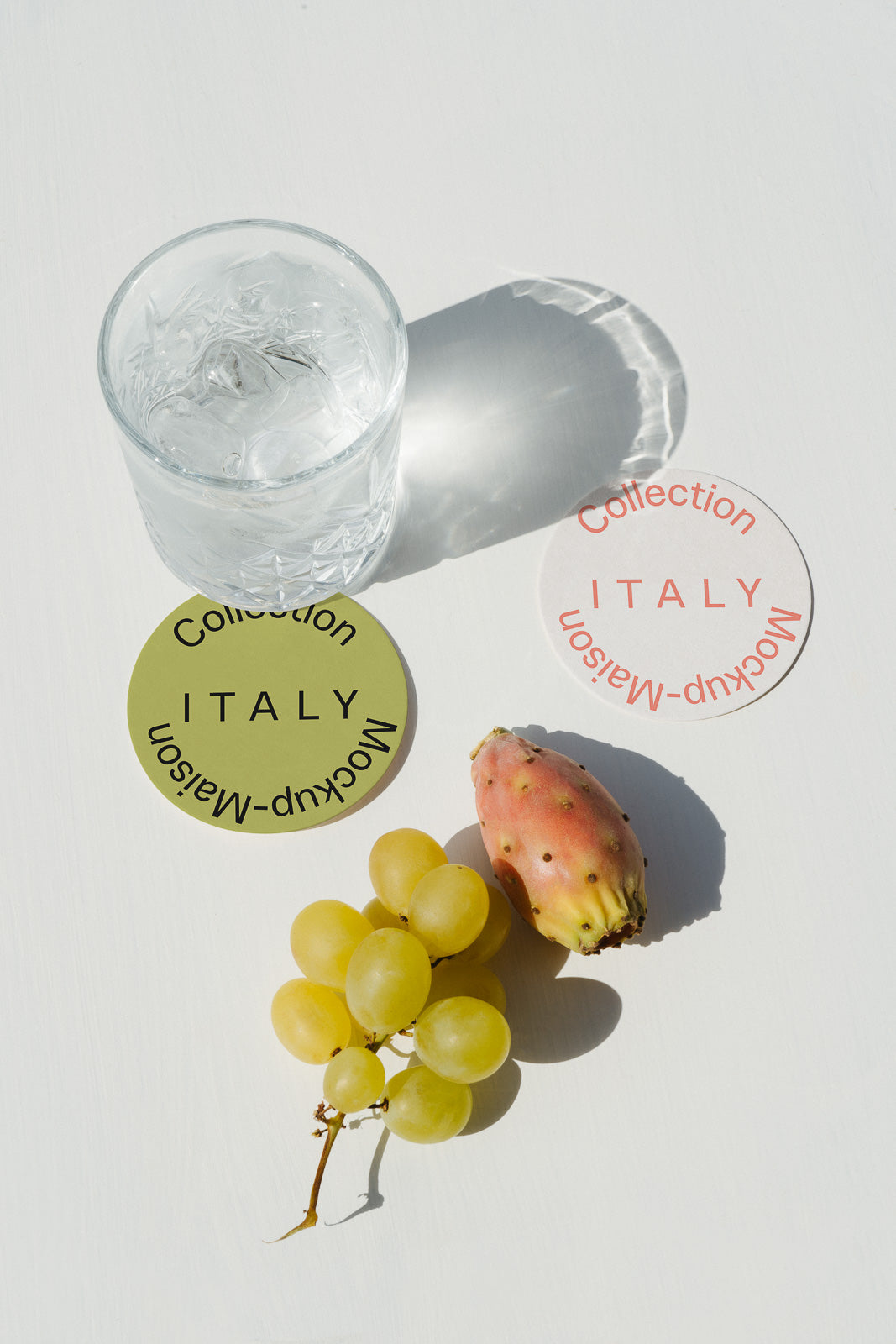 PSD mockup of a glass with ice casting a shadow near two circular coasters labeled "Collection Italy Mockup-Maison," with grapes and a prickly pear.
