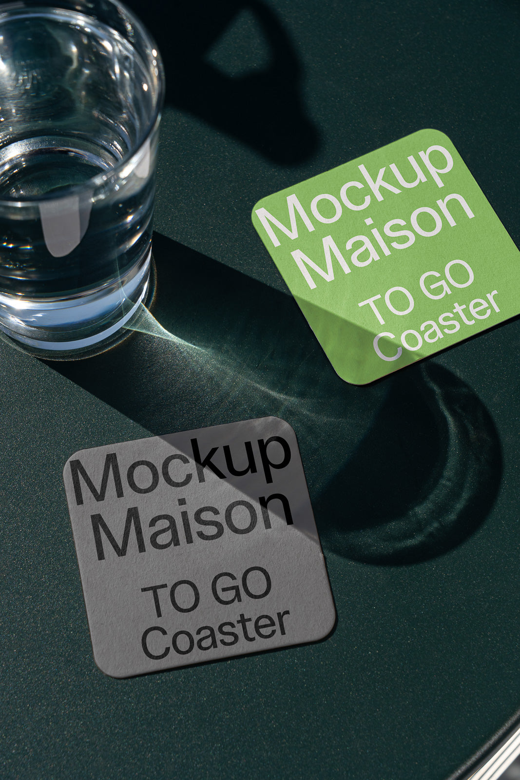 PSD mockup of two square coasters on a dark surface, one green and one gray, with sunlight reflecting through a nearby glass of water.