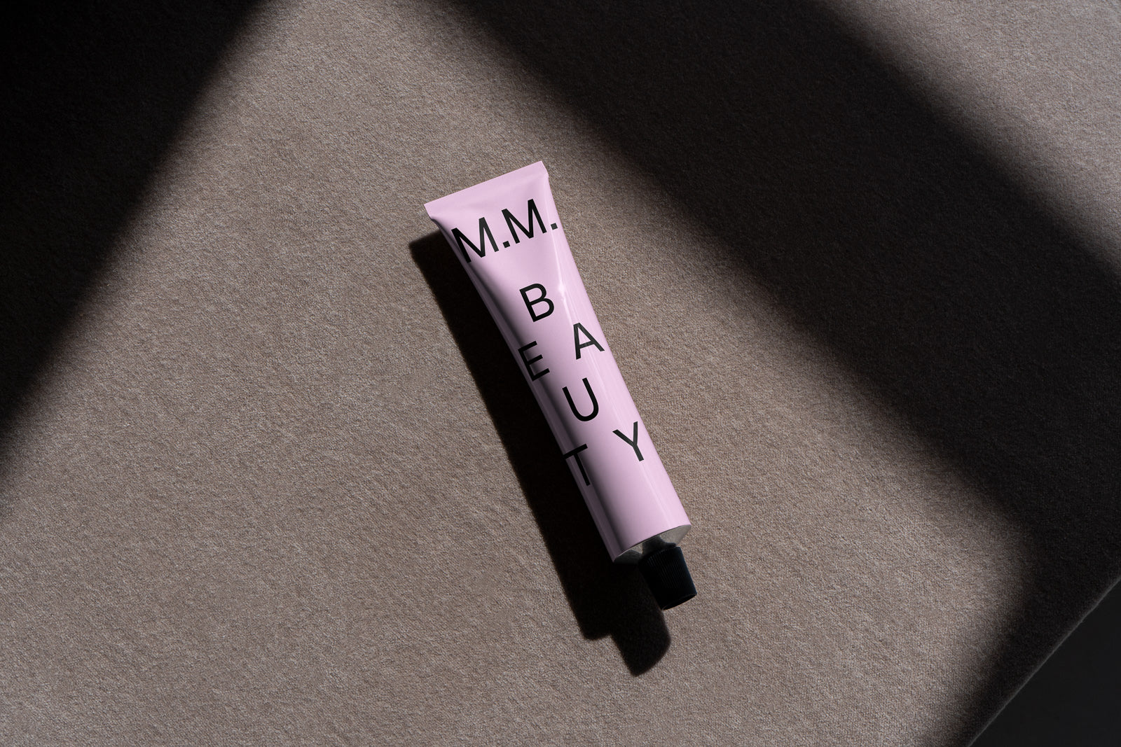PSD mockup of a pink cosmetic tube labeled "M.M. Beauty" placed on a textured surface, casting a shadow in soft natural light.