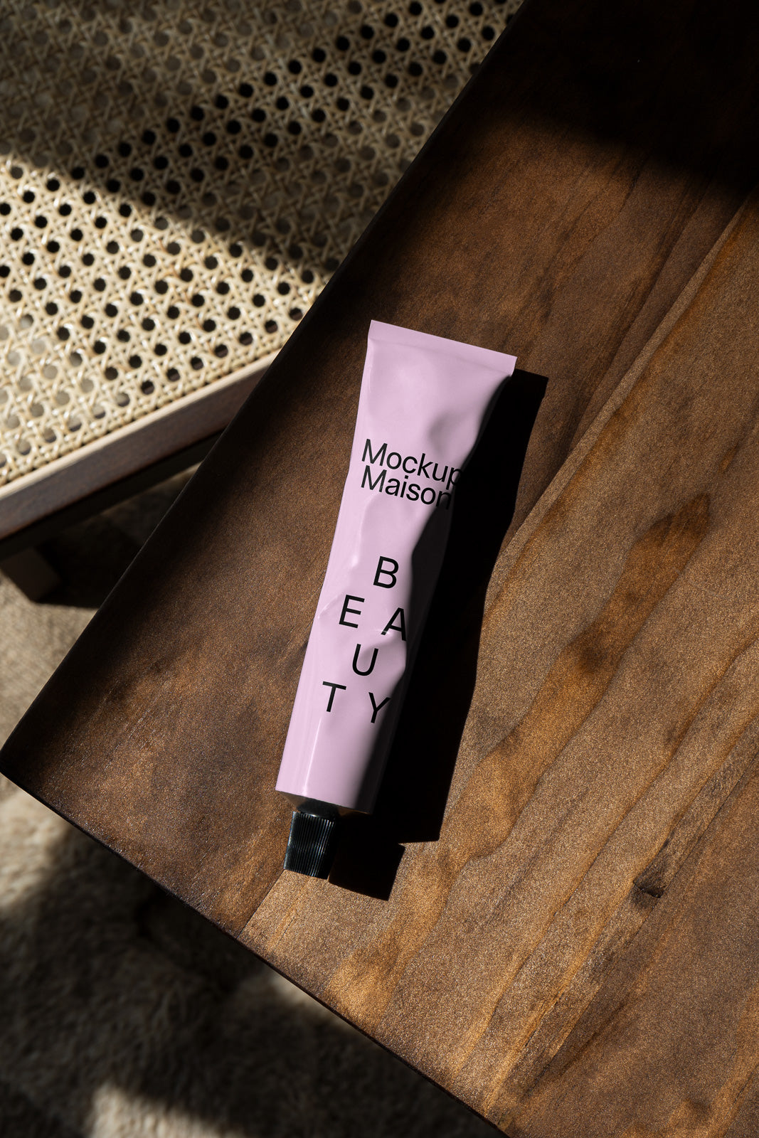PSD mockup of a pink beauty product tube labeled "Mockup Maison Beauty" on a wooden surface with woven textures and soft lighting.