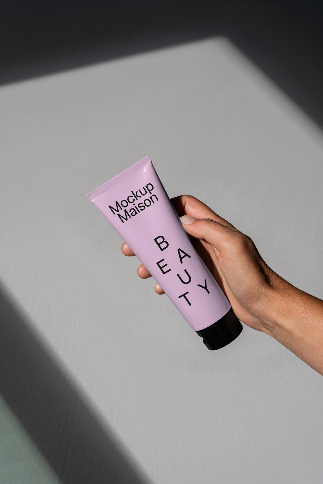 PSD mockup of a hand holding a lavender-colored beauty tube labeled "Mockup Maison" and "BEAUTY," casting a shadow on a gray background.