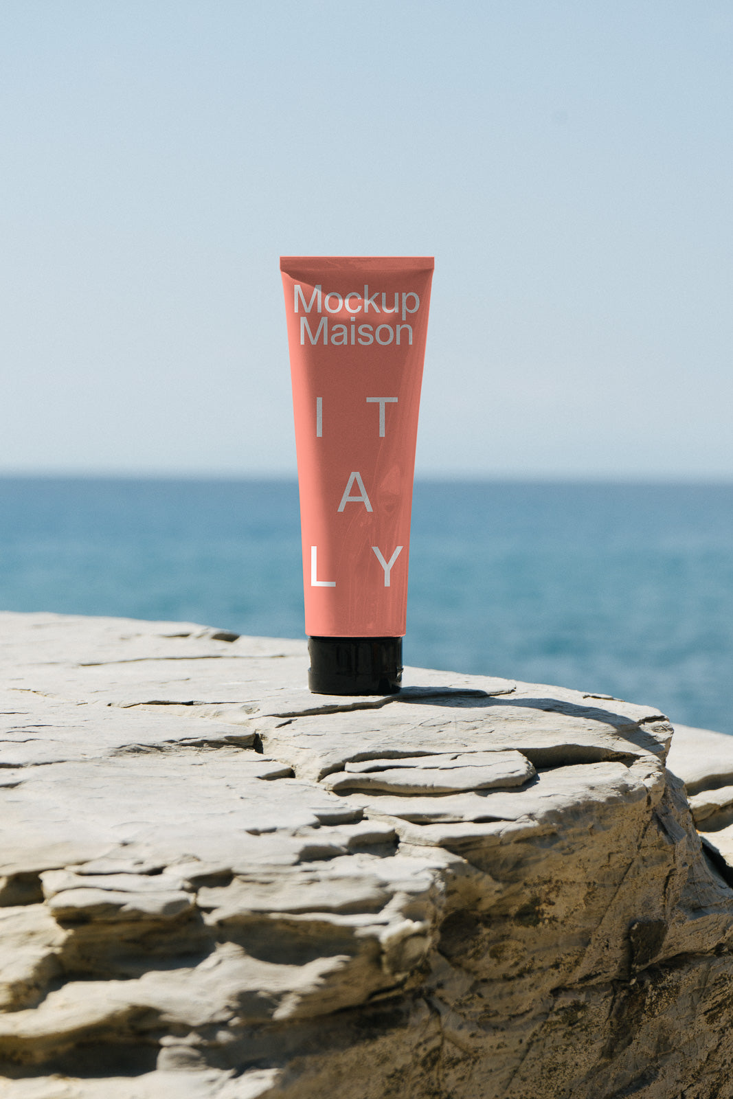 PSD mockup of a coral-colored tube with "Mockup Maison Italy" text, standing on a rocky surface against a backdrop of the sea and clear sky.