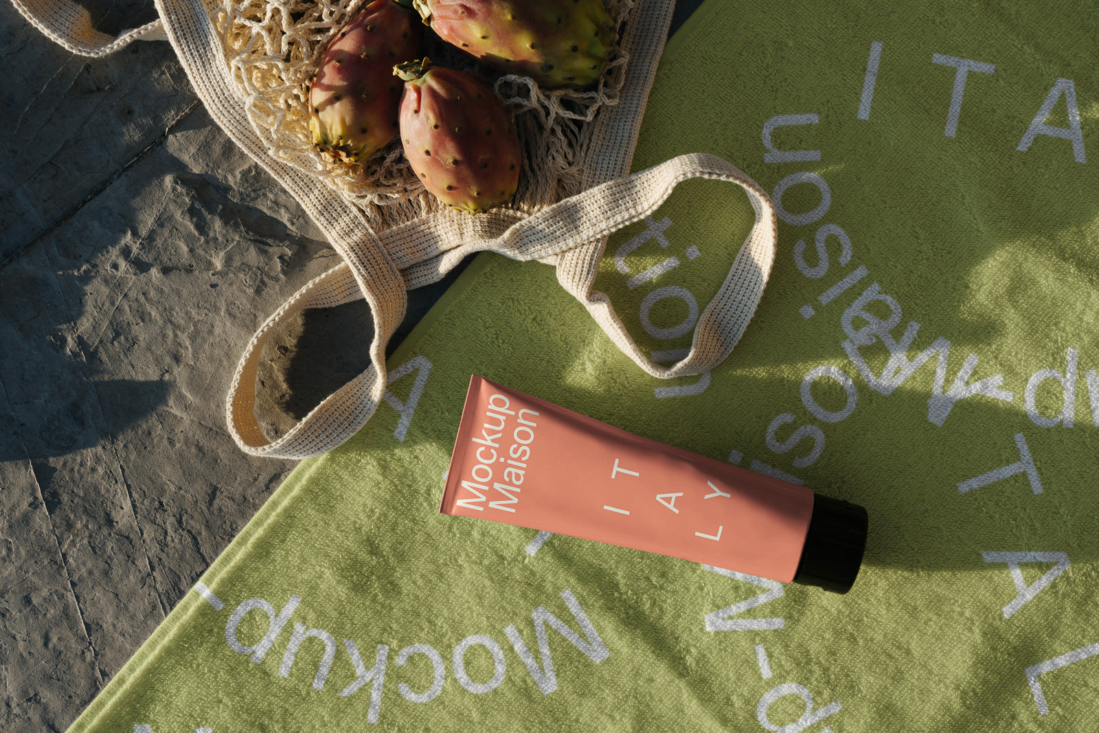 PSD mockup of a tube labeled "Mockup Maison Italy" resting on a green towel with a reusable bag containing prickly pears nearby.