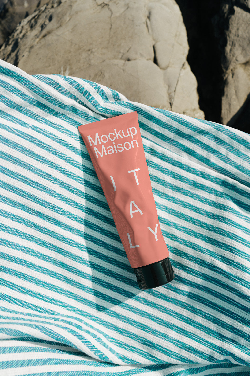 PSD mockup of a pink tube labeled "Mockup Maison ITALY" resting on a turquoise and white striped fabric, set against a rocky background.