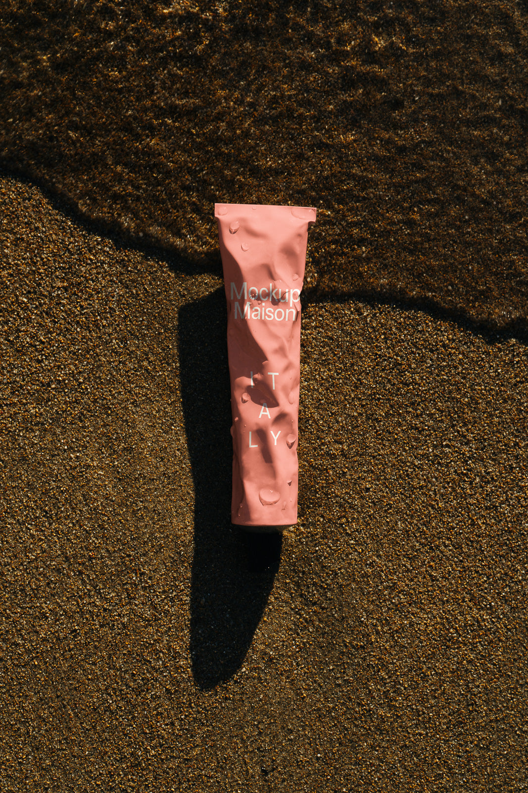 PSD mockup of a pink cosmetic tube with water droplets laying on wet sand, casting a shadow in the sunlight.