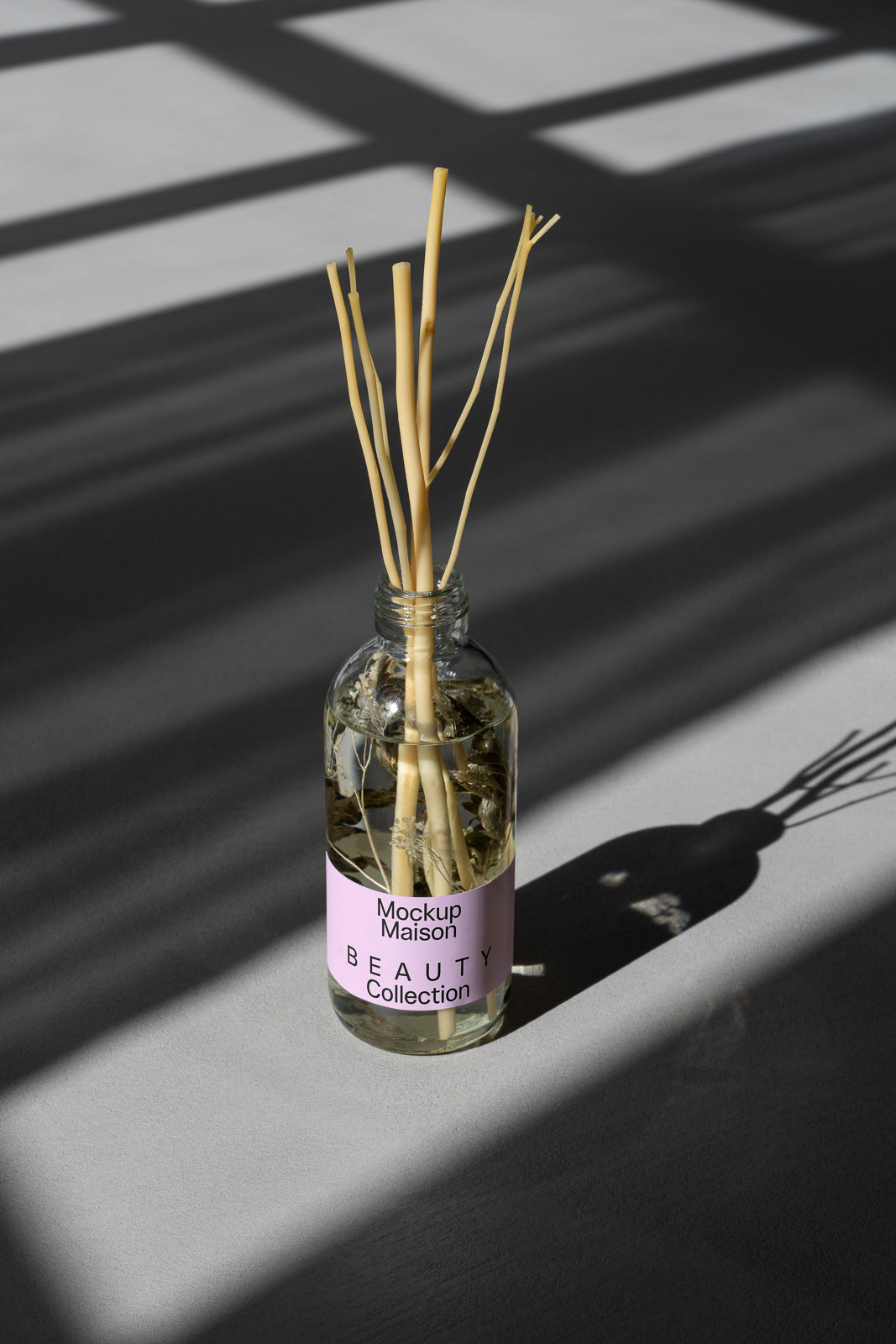 PSD mockup of a glass bottle with diffuser sticks labeled "Maison Beauty Collection," casting shadows on a gray surface with geometric light patterns.