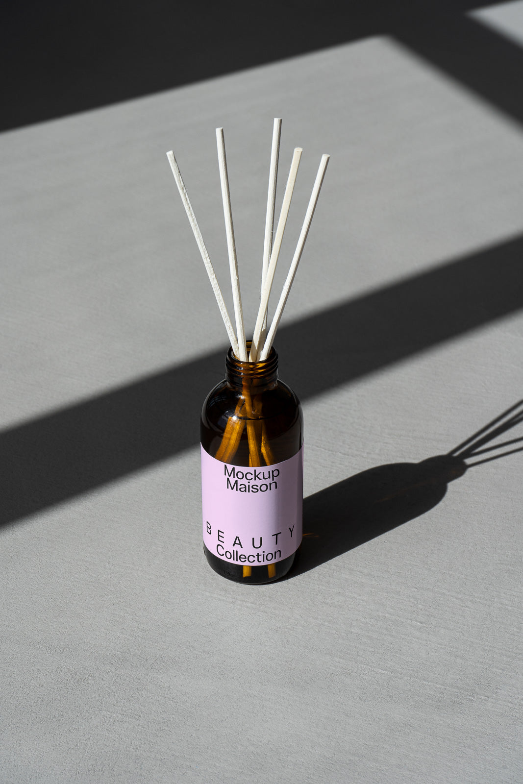PSD mockup of a brown bottle with a pink label titled “Mockup Maison Beauty Collection” containing reed sticks, casting shadows on a gray surface.