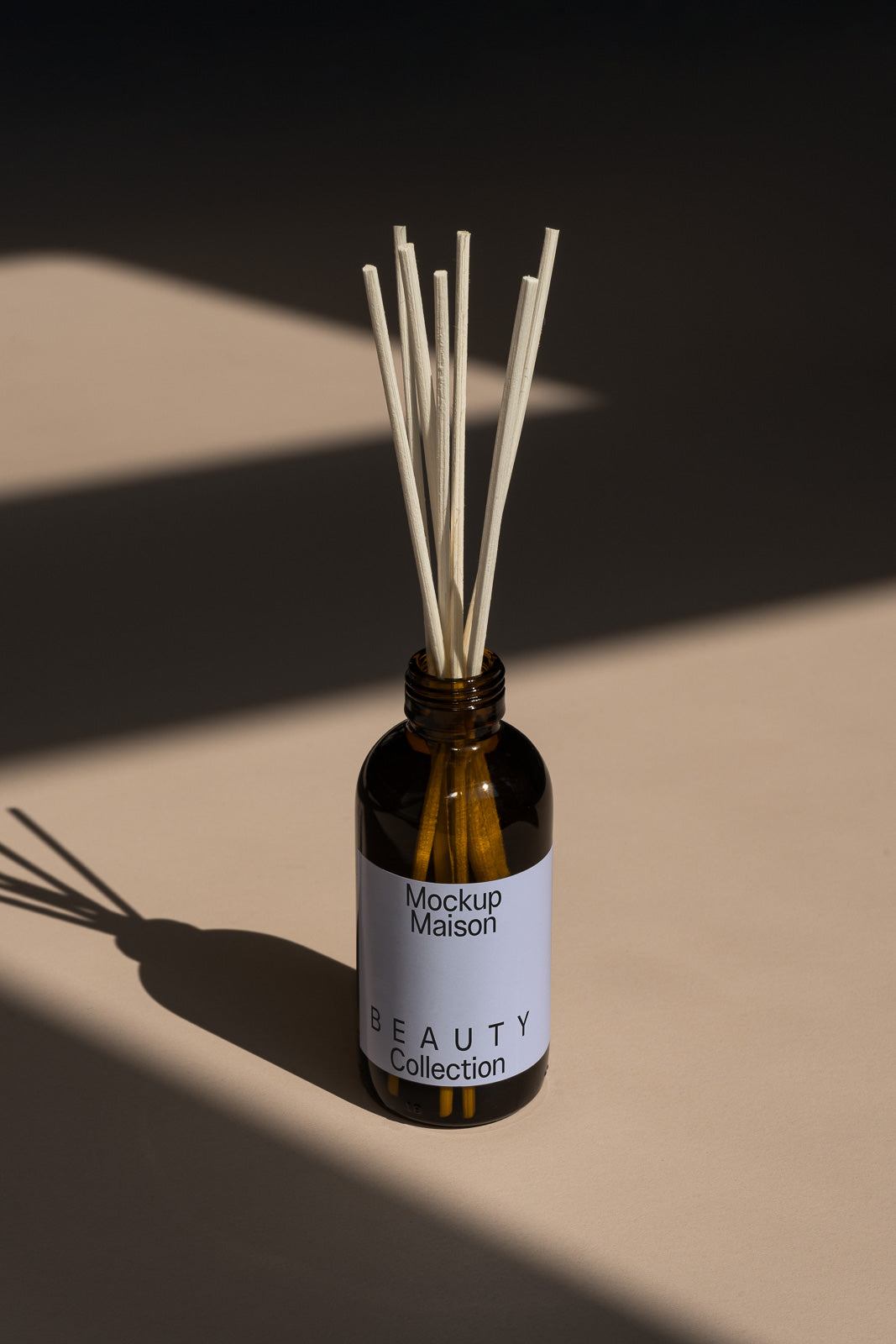 PSD mockup of a brown bottle with reeds, labeled "Mockup Maison Beauty Collection," casting a shadow on a beige surface.