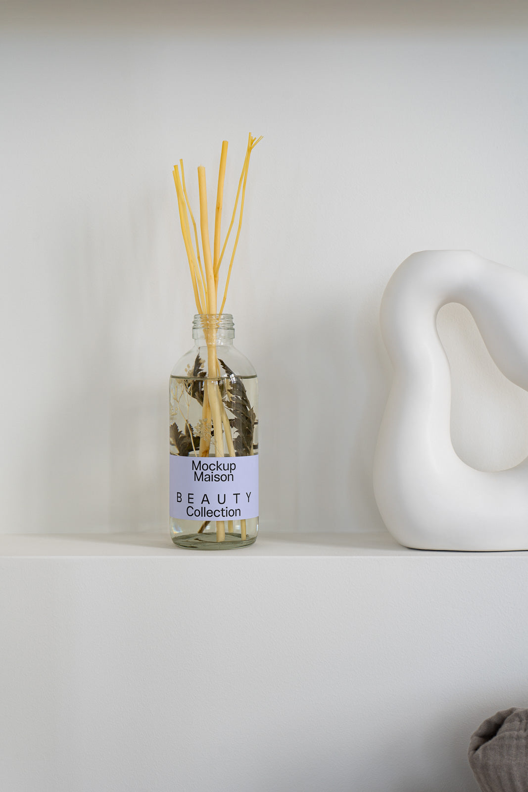 PSD mockup of a glass bottle labeled "Mockup Maison Beauty Collection" with aroma sticks, placed on a shelf next to a white abstract sculpture.