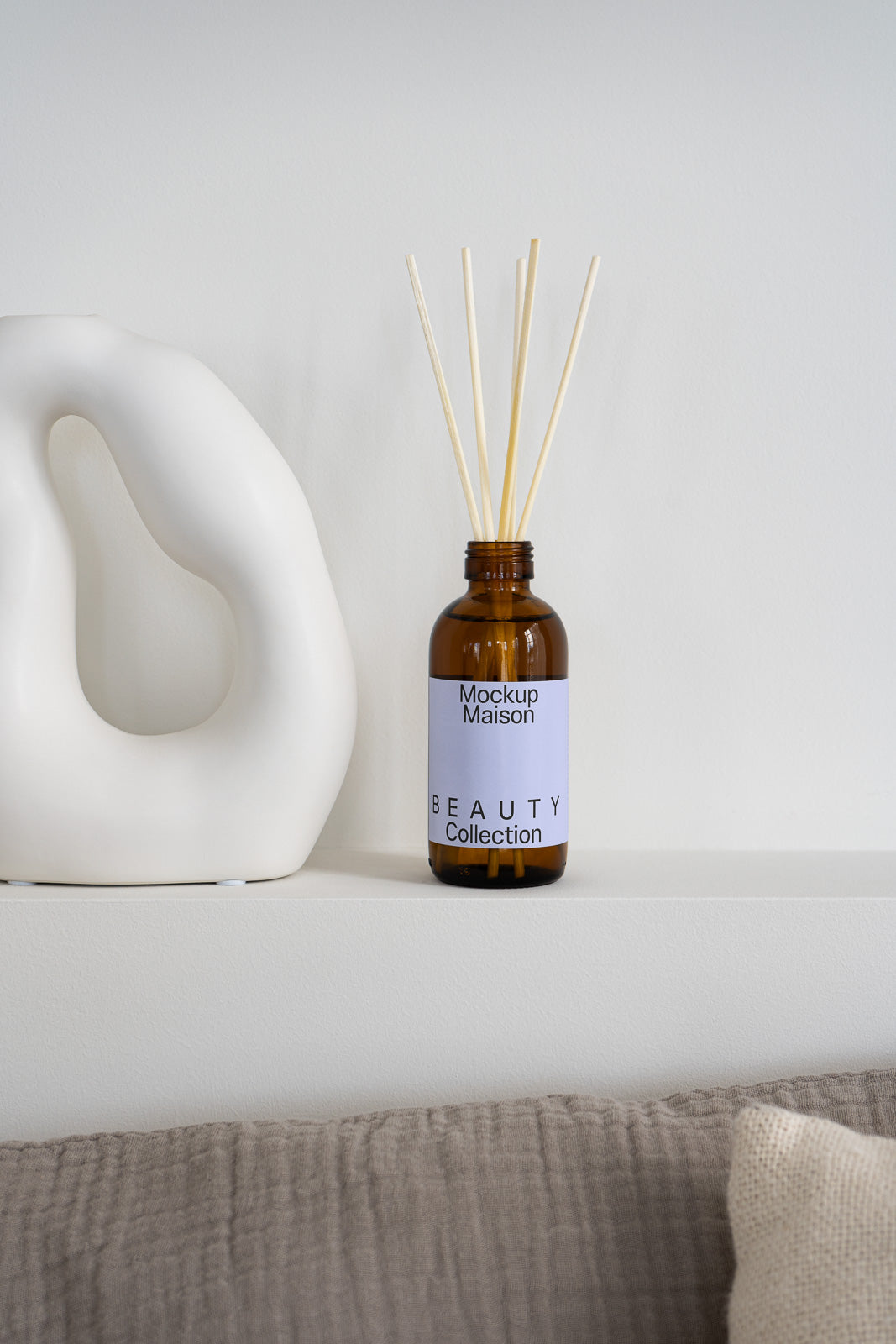 PSD mockup of a brown glass reed diffuser bottle labeled "Mockup Maison Beauty Collection" next to a white ceramic sculpture on a white shelf.
