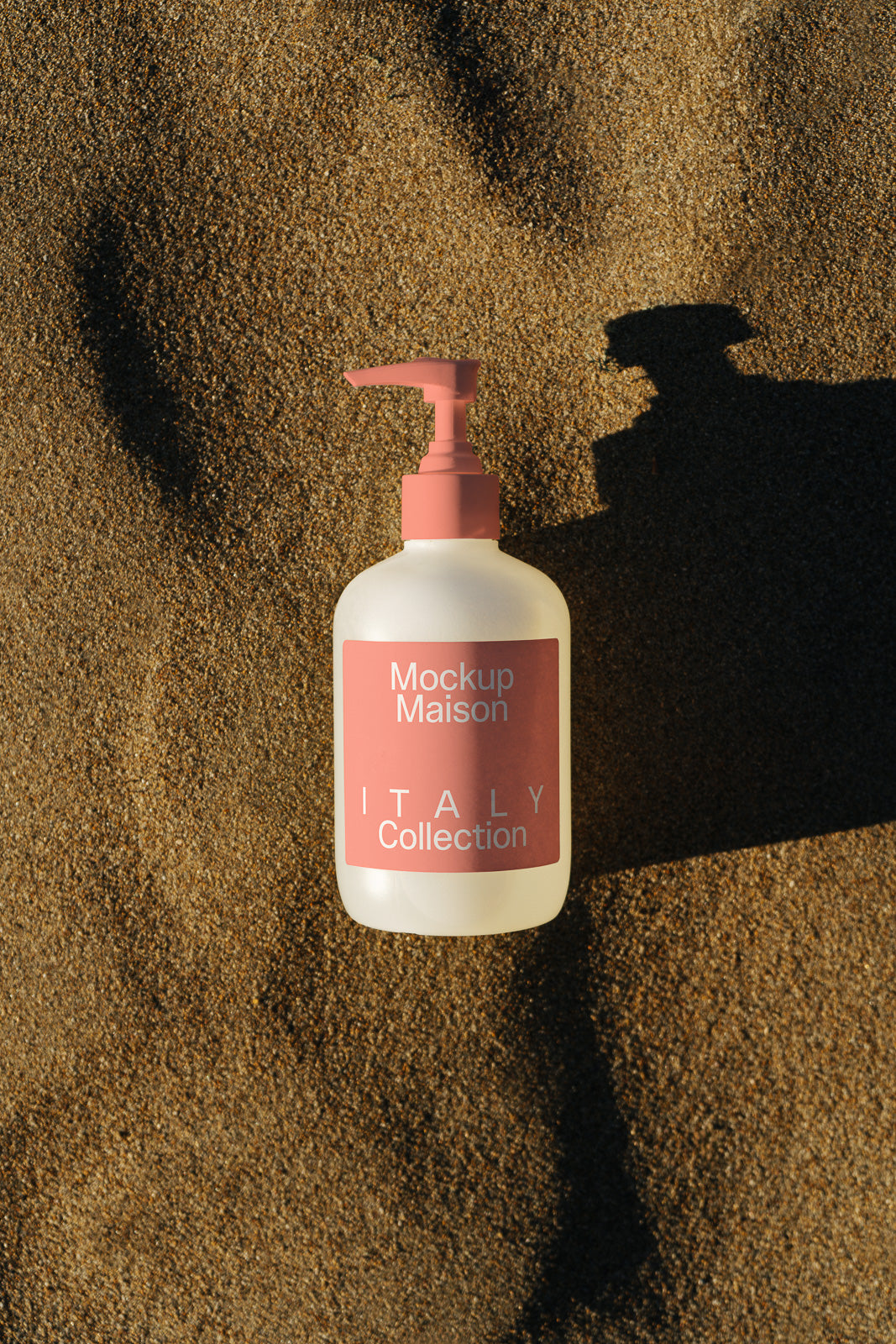 PSD mockup of a bottle with a pink pump cap labeled "Mockup Maison Italy Collection" resting on textured sand casting a shadow.