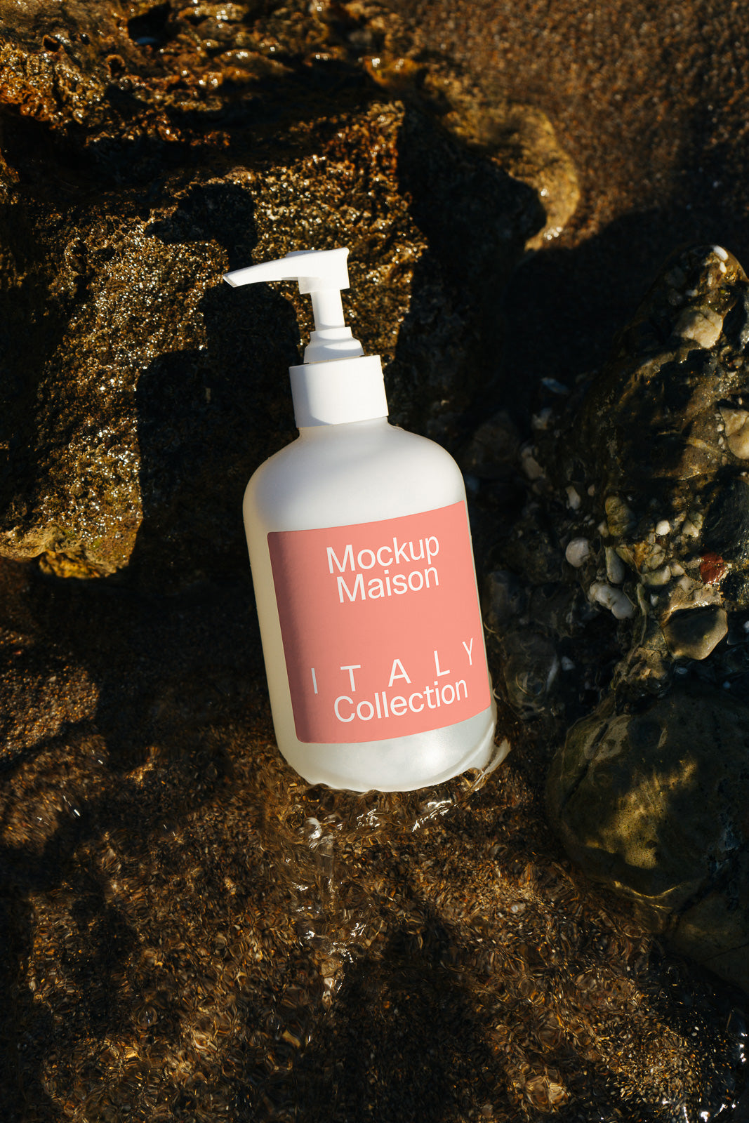 PSD mockup of a white pump bottle labeled "Mockup Maison Italy Collection," placed among wet rocks with shallow water surrounding it.