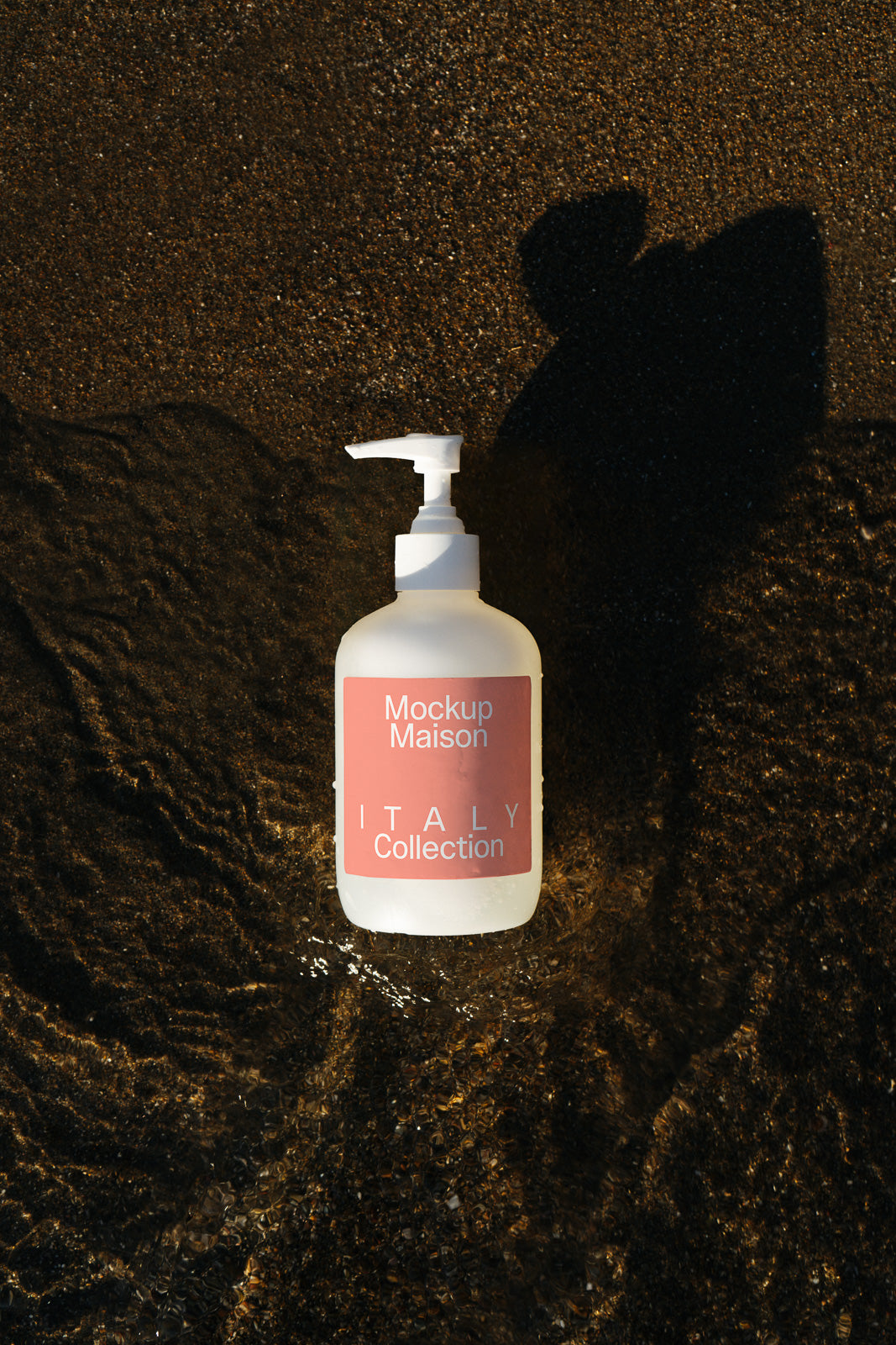PSD mockup of a frosted pump bottle labeled "Mockup Maison Italy Collection" placed on dark, wet sand with water ripples and shadow.