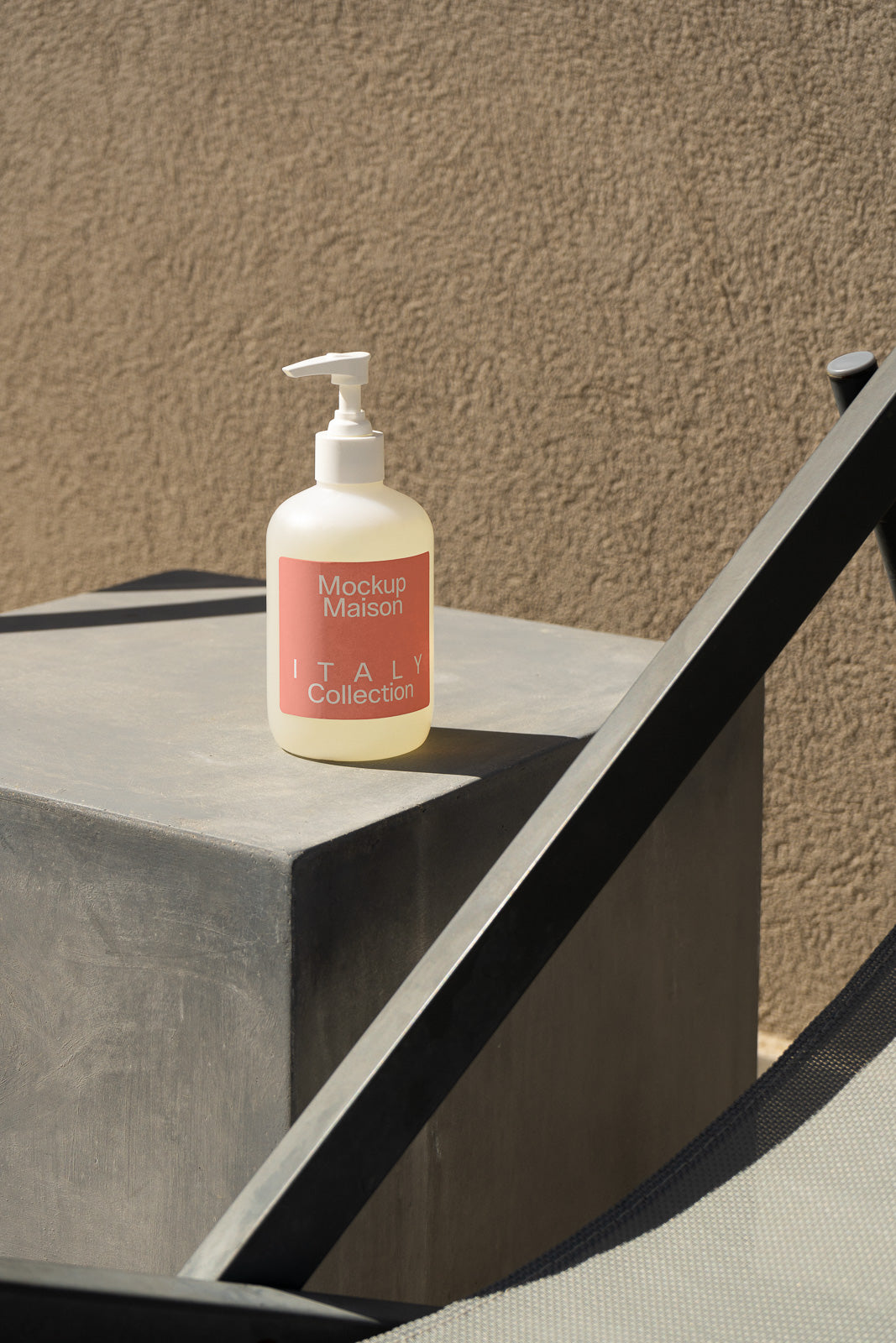 PSD mockup of a pump bottle labeled "Mockup Maison Italy Collection" on a concrete surface, next to modern metal furniture, with a textured wall background.