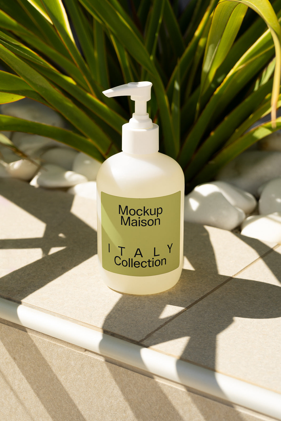 PSD mockup of a frosted pump bottle labeled "Mockup Maison ITALY Collection" set on a tiled surface with green plant leaves and white stones in the background.