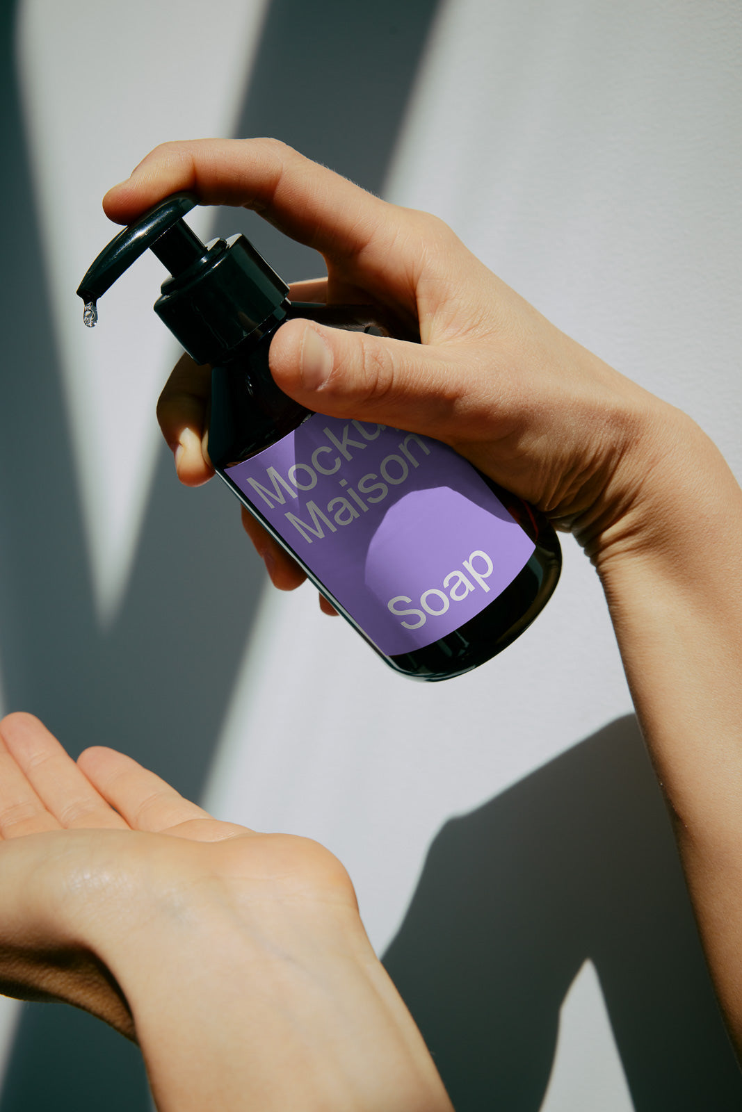 PSD mockup of a hand pressing a pump bottle labeled "Mockup Maison Soap" with a droplet falling onto another hand.