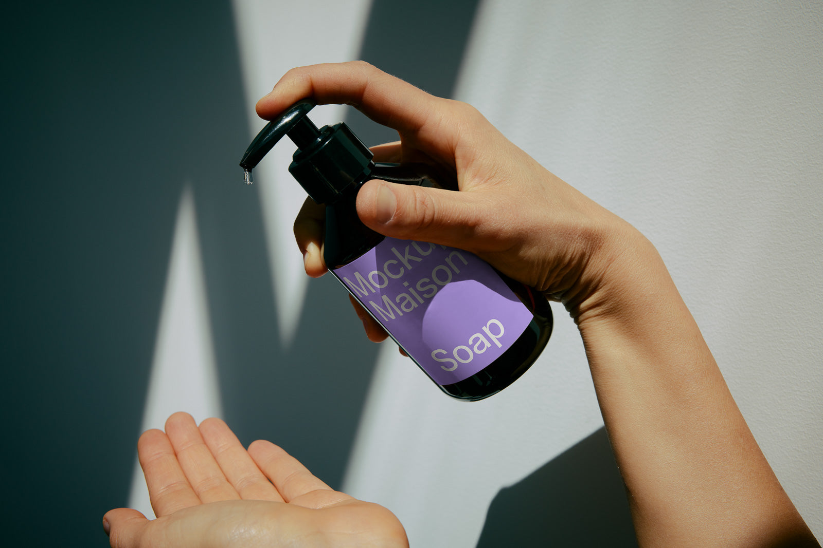 PSD mockup of a person dispensing soap from a dark bottle with a purple label reading "Maison Mockup Soap" onto their palm, set against a softly lit wall.