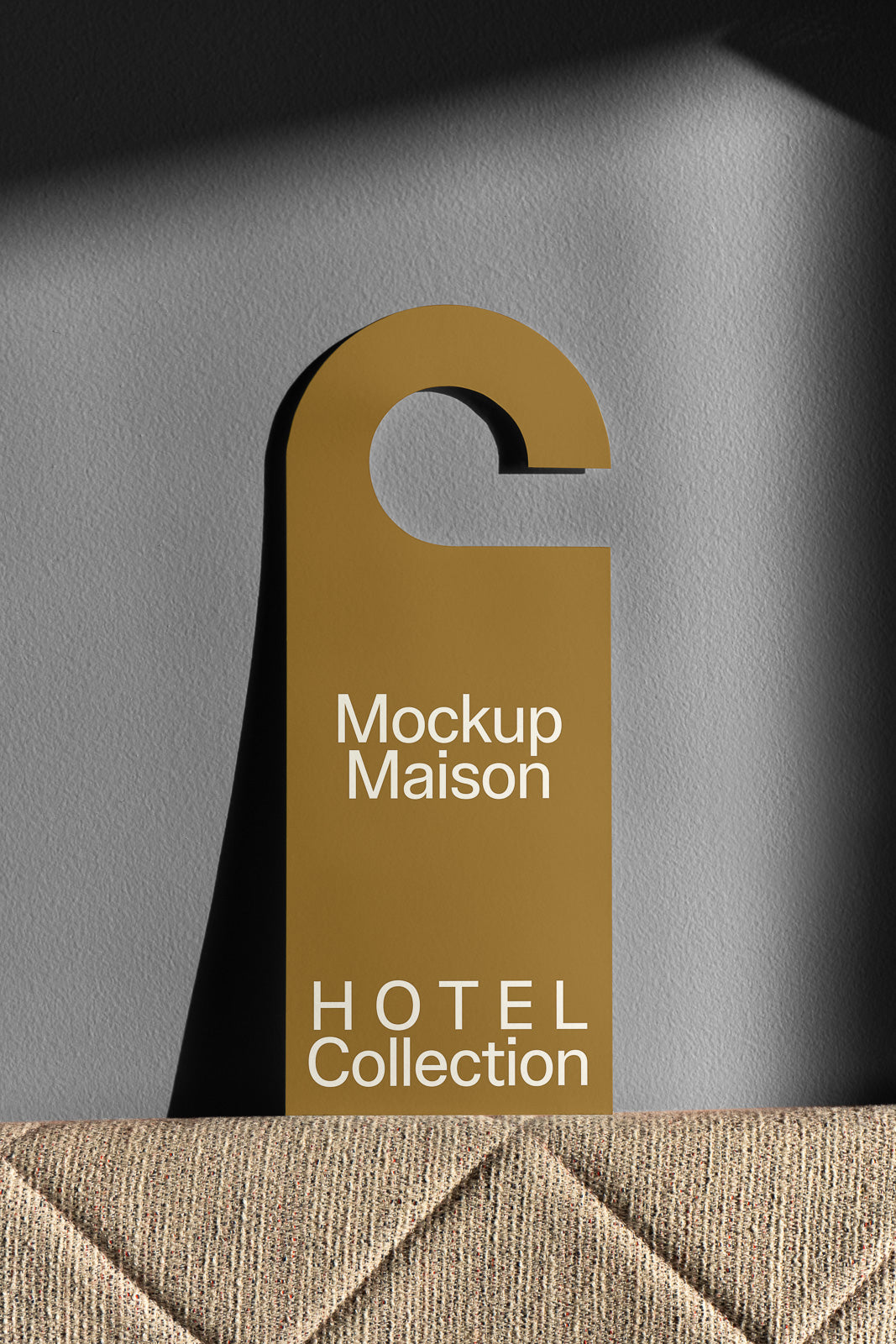 PSD mockup of a gold hotel door hanger with "Mockup Maison" and "HOTEL Collection" printed on it, leaning against a textured fabric background.