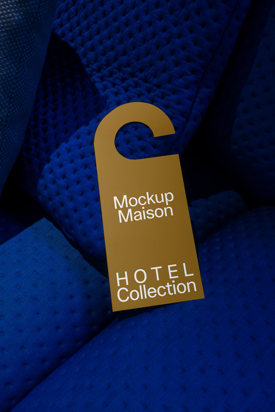 PSD mockup of a gold door hanger labeled "Mockup Maison HOTEL Collection" resting on a textured blue surface.