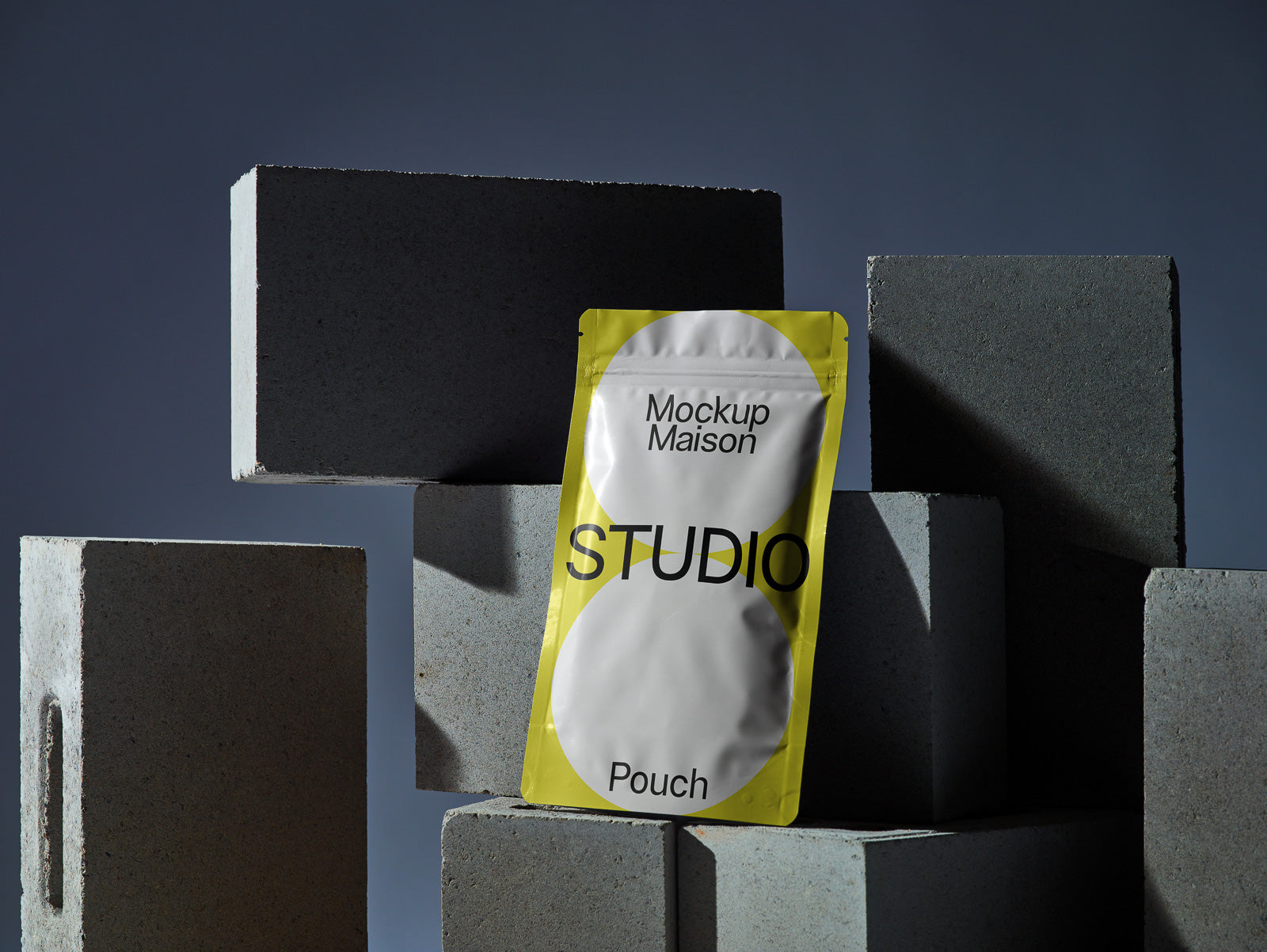 PSD mockup of a yellow stand-up pouch with "Mockup Maison" and "Studio Pouch" text, positioned among gray concrete blocks against a dark background.