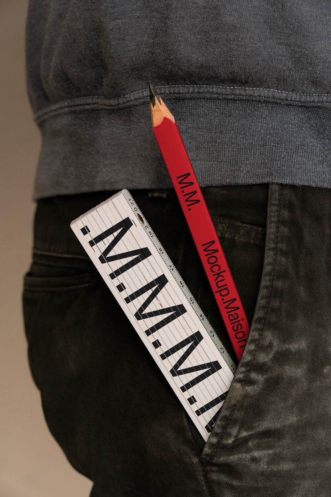 PSD mockup of a large red pencil and a ruler with "M.M." in a jeans pocket, against a textured grey background.