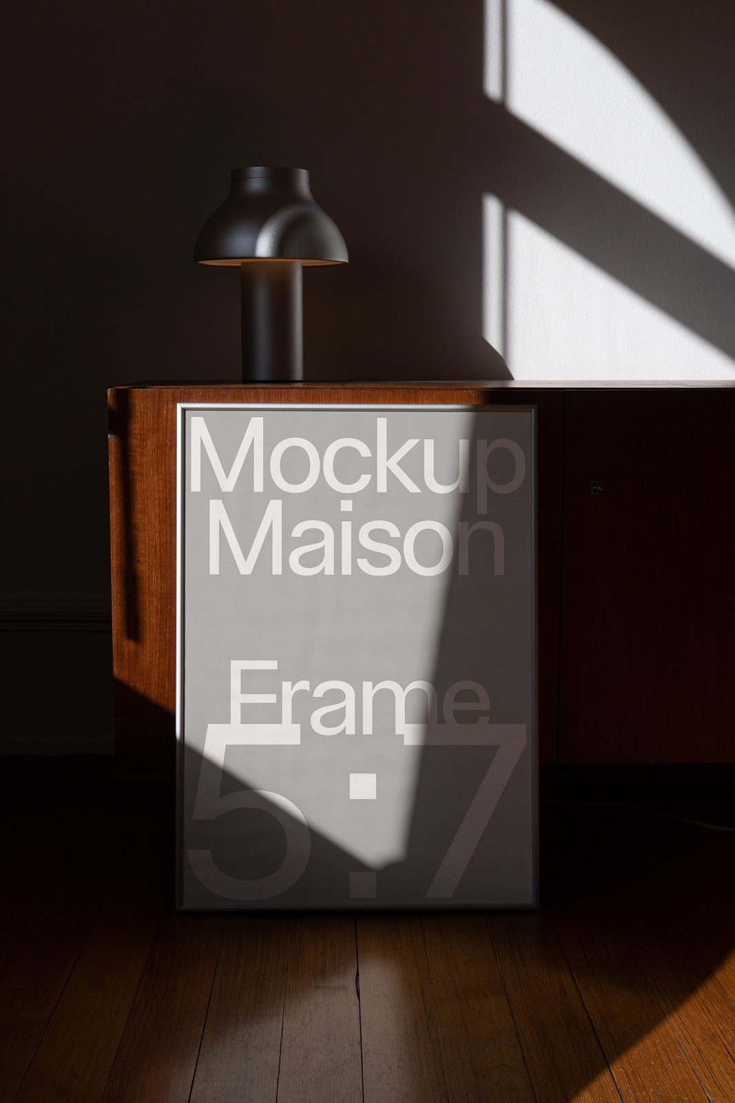 PSD mockup of a framed poster leaning against a wooden cabinet, with a modern lamp on top, lit by natural sunlight casting shadows.