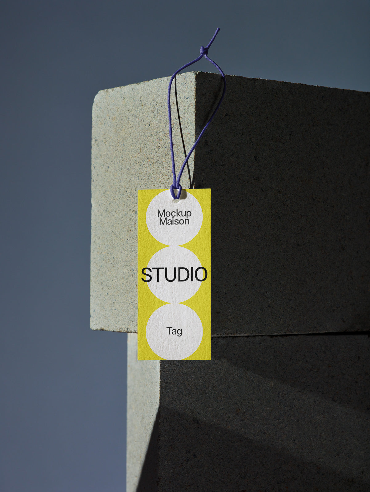 PSD mockup of a yellow rectangular tag with a purple string, labeled "Mockup Maison Studio Tag," hanging on a gray concrete block corner.