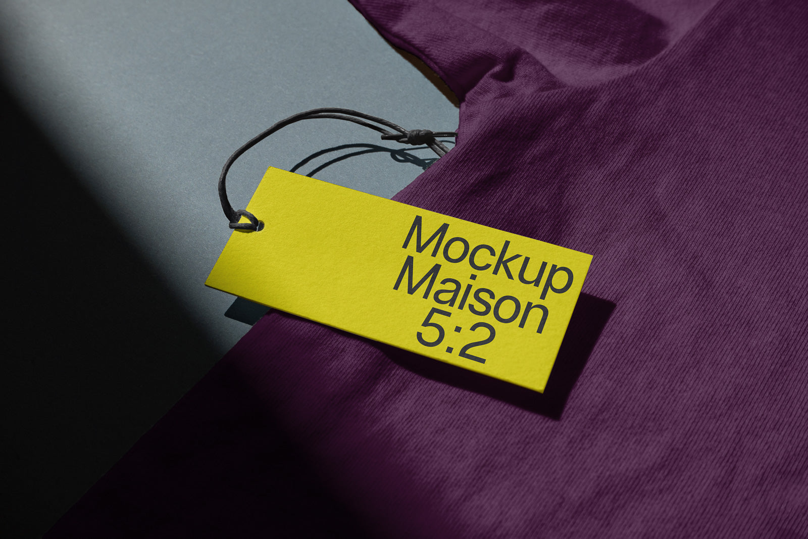 PSD mockup of a yellow tag labeled "Mockup Maison 5:2" attached to a purple fabric with a dark string, partially illuminated by light.