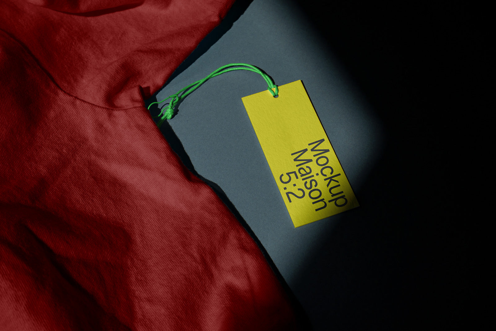 PSD mockup of a yellow tag with green string attached to a dark surface, partially covered by red fabric. The tag reads "Mockup Maison 5:2".
