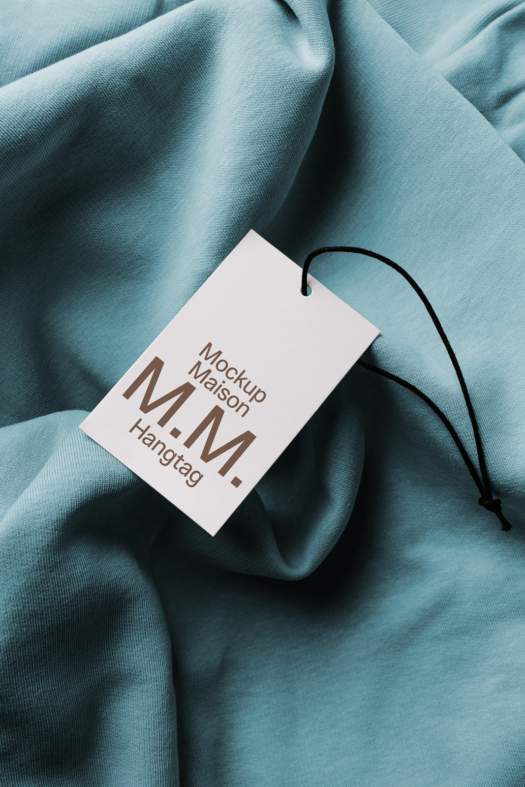 PSD mockup of a hangtag labeled "Mockup Maison M.M." resting on blue fabric with a black string attached, showcasing product branding potential.