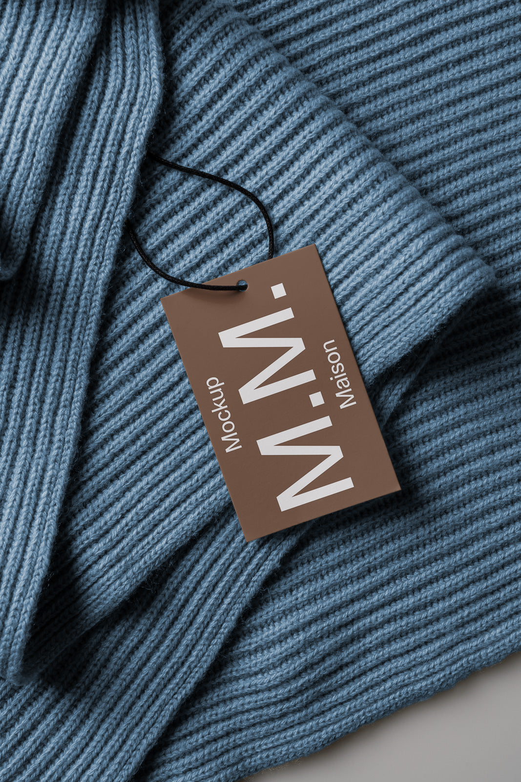PSD mockup of a brown tag labeled "M.N.- Maison" with a black string, placed on a textured blue knit fabric.
