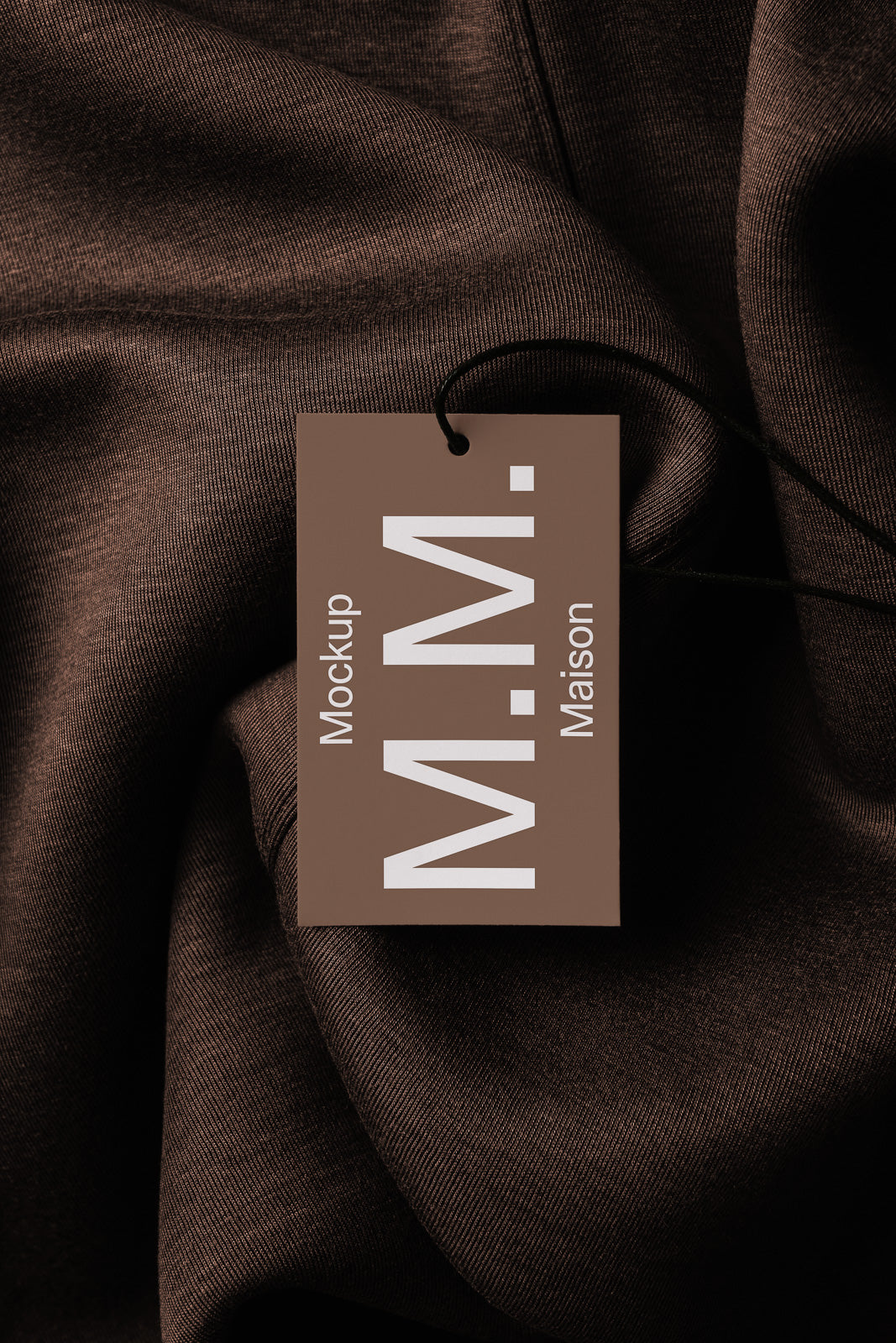 PSD mockup of a brown tag labeled "Mockup Maison M.M." lying on dark textured fabric.