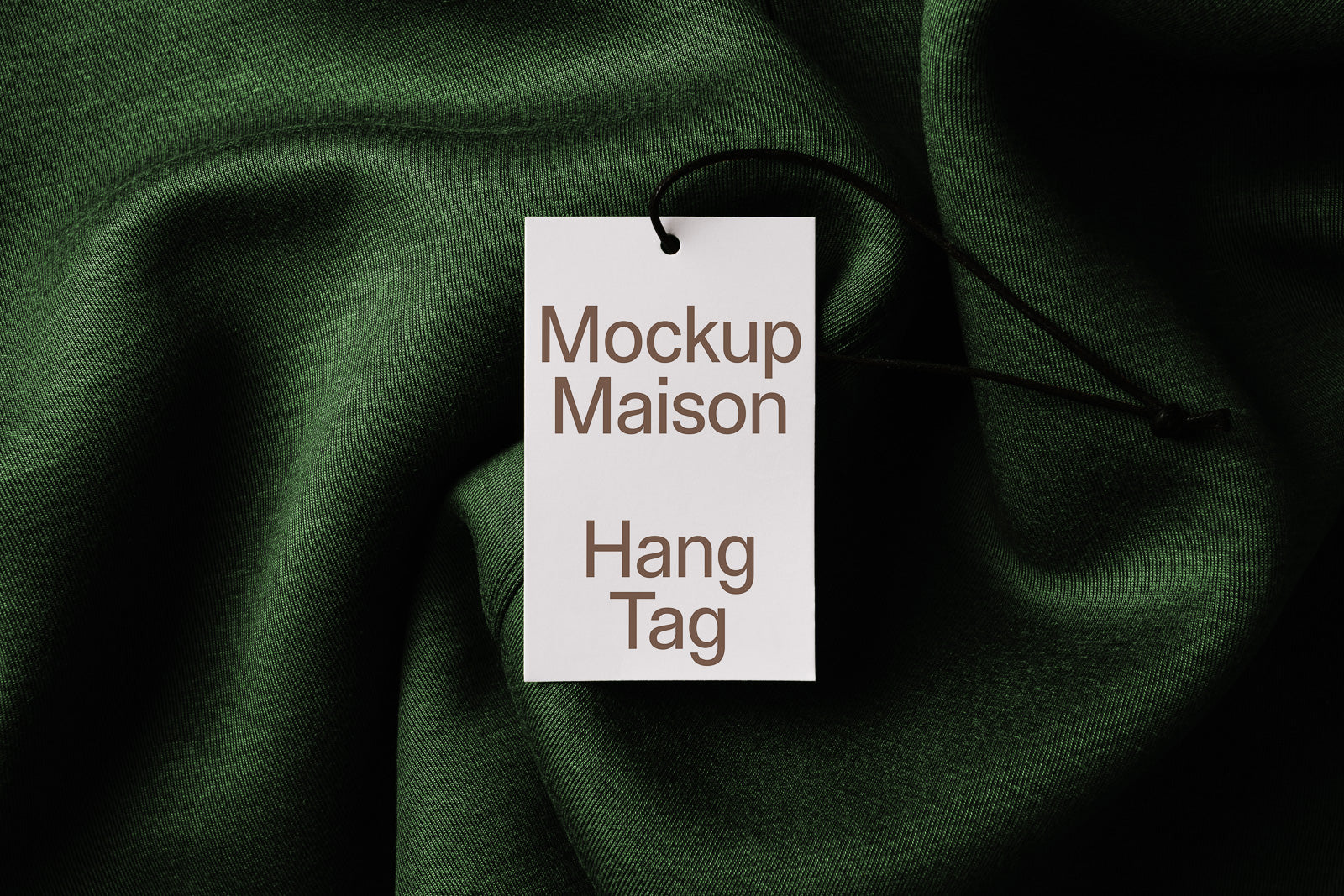 PSD mockup of a hang tag with "Mockup Maison Hang Tag" text, lying on textured dark green fabric with a black string attached.