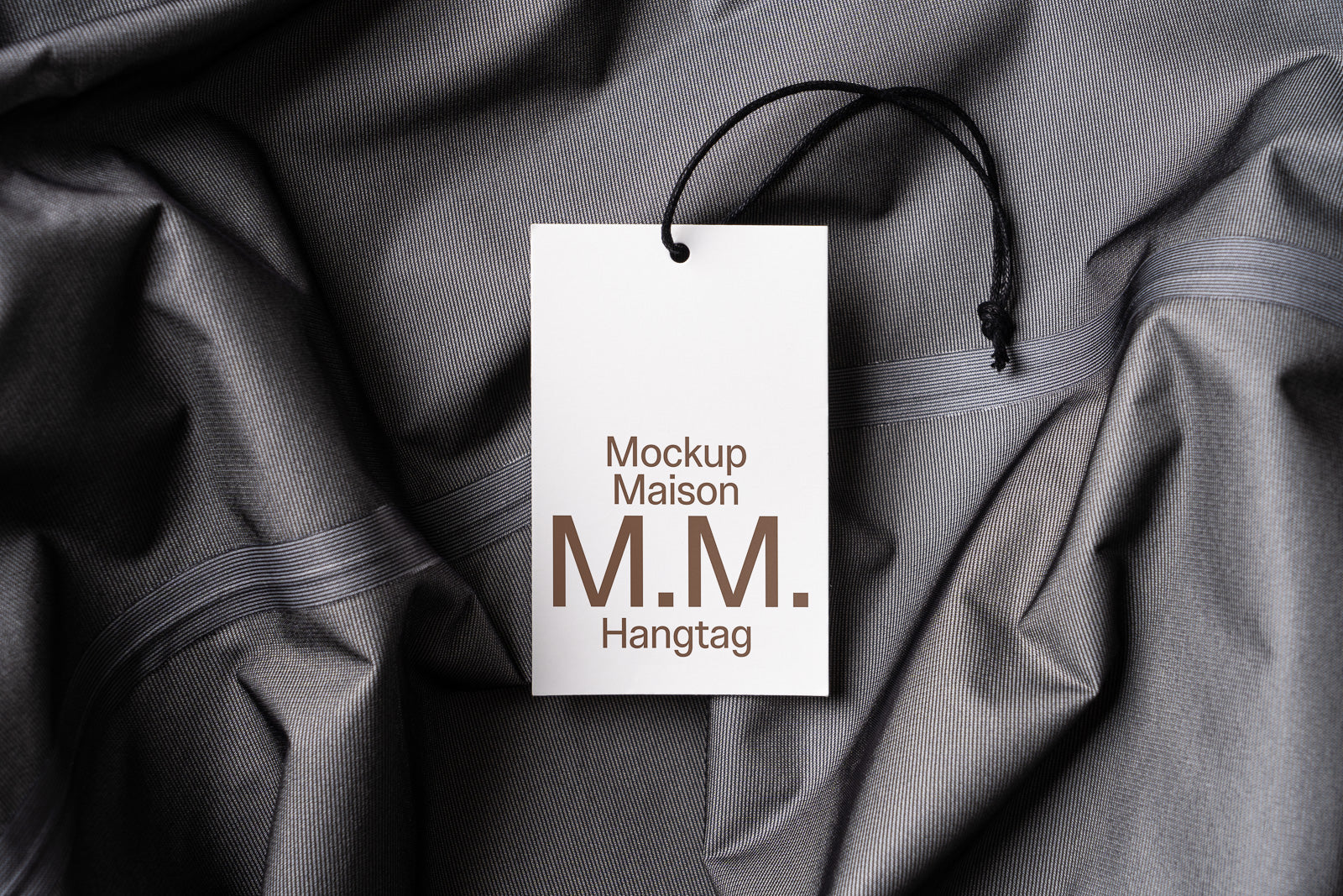 PSD mockup of a clothing hangtag with "Mockup Maison M.M." printed, lying on a grey fabric background.
