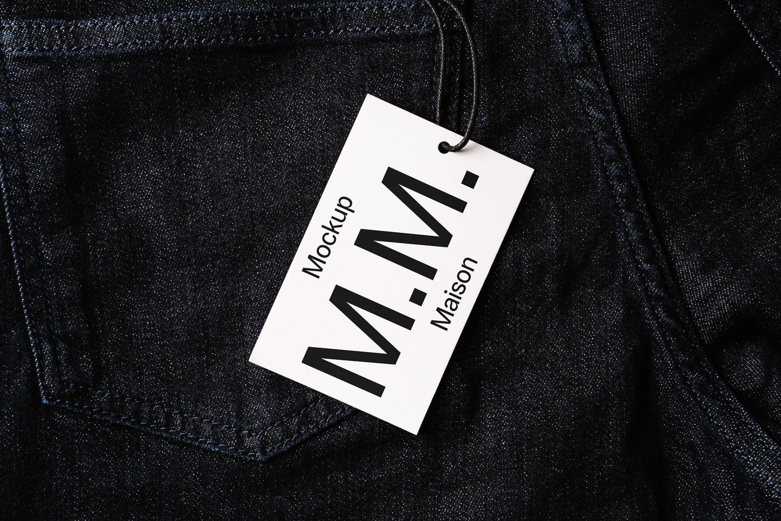 PSD mockup of a white clothing tag labeled "Mockup M.M. Maison" attached to dark denim fabric, displaying branding elements and material texture.