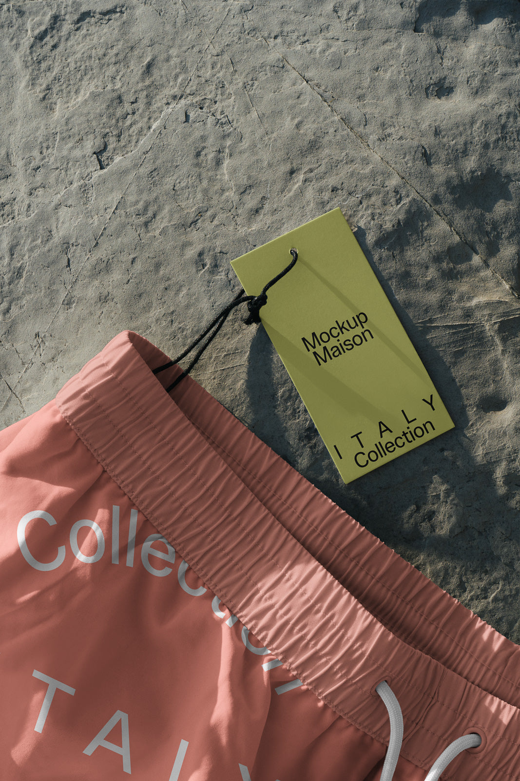 PSD mockup of pink swim trunks with a drawstring waistband and an attached green tag labeled "Mockup Maison, ITALY Collection" placed on a textured surface.
