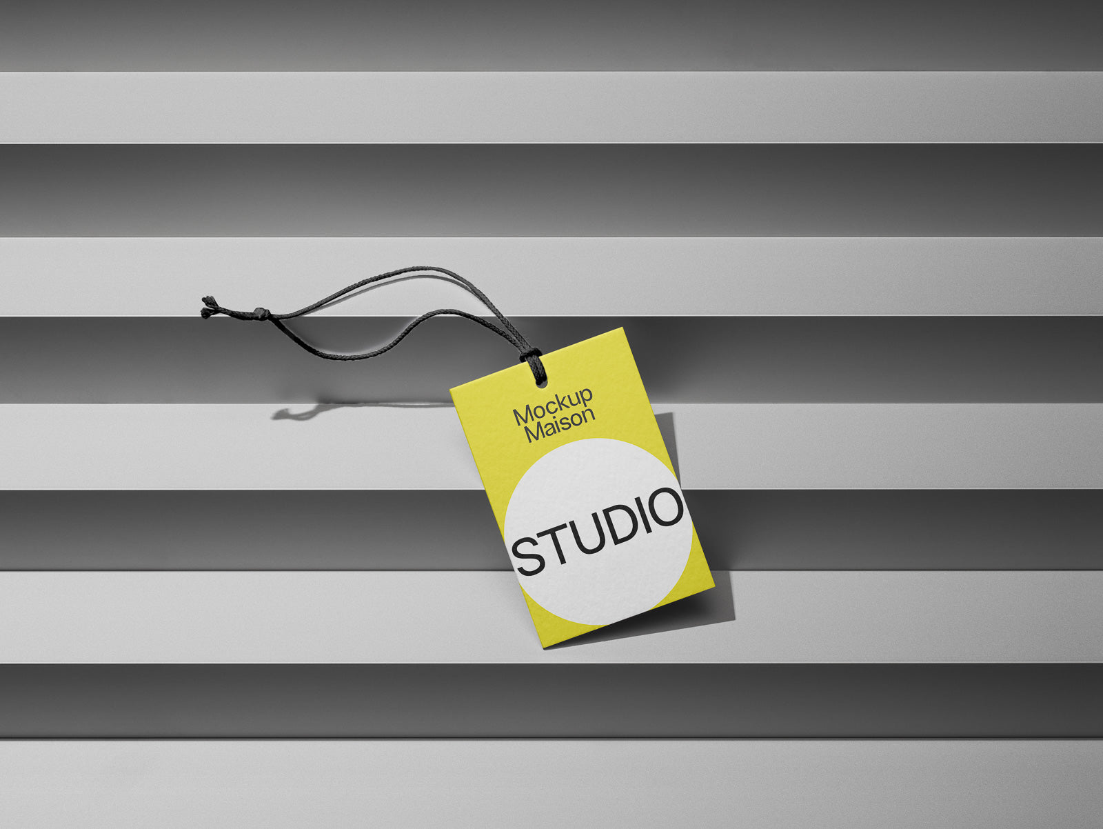 PSD mockup of a yellow and white clothing tag labeled "Mockup Maison STUDIO" on a series of horizontal gray blinds.