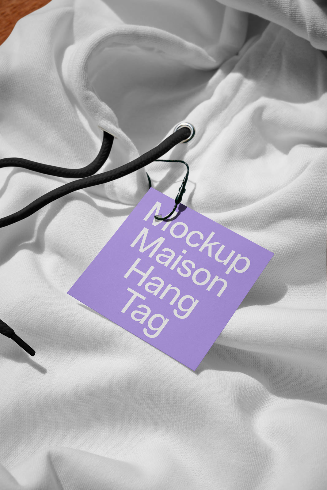 PSD mockup of a purple rectangular hang tag attached to a white hoodie with black drawstrings, lying on a wooden surface.
