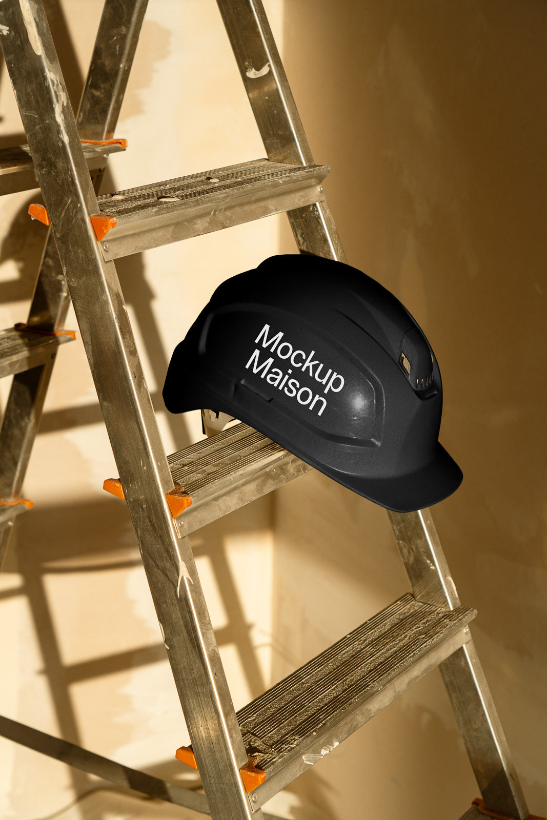 PSD mockup of a black hard hat labeled "Mockup Maison" positioned on a metal ladder with orange accents, against a beige background.