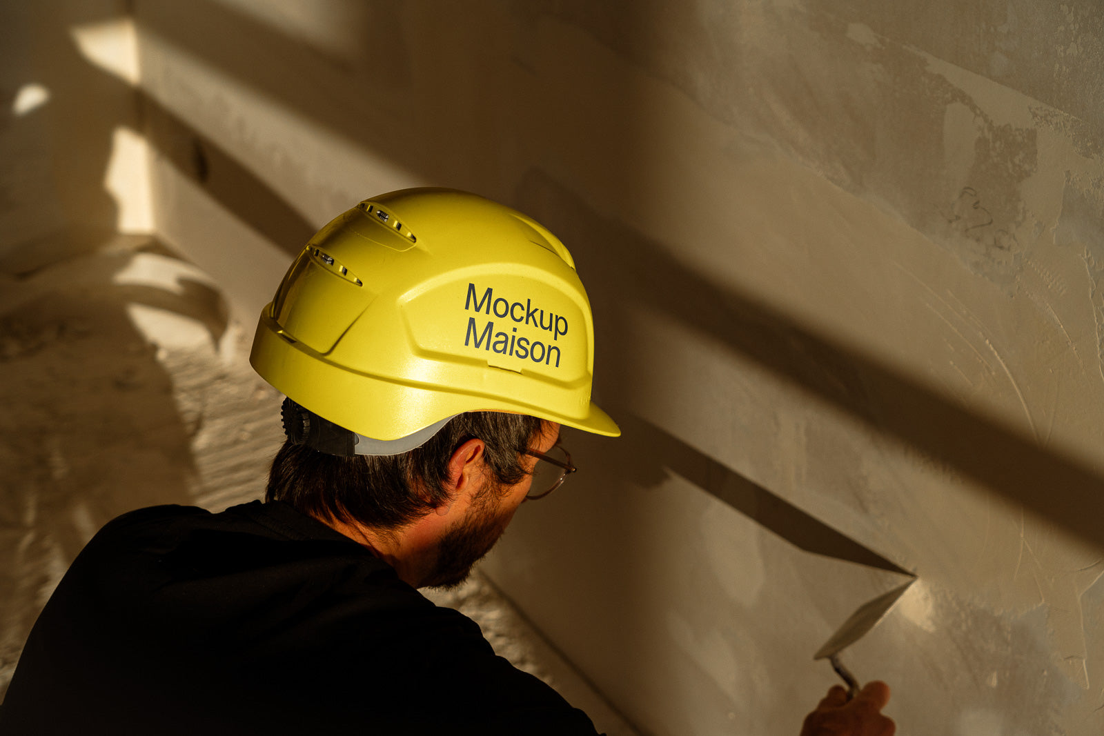 PSD mockup of a construction worker wearing a yellow hard hat with "Mockup Maison" text, applying plaster to a wall with a trowel in low light.