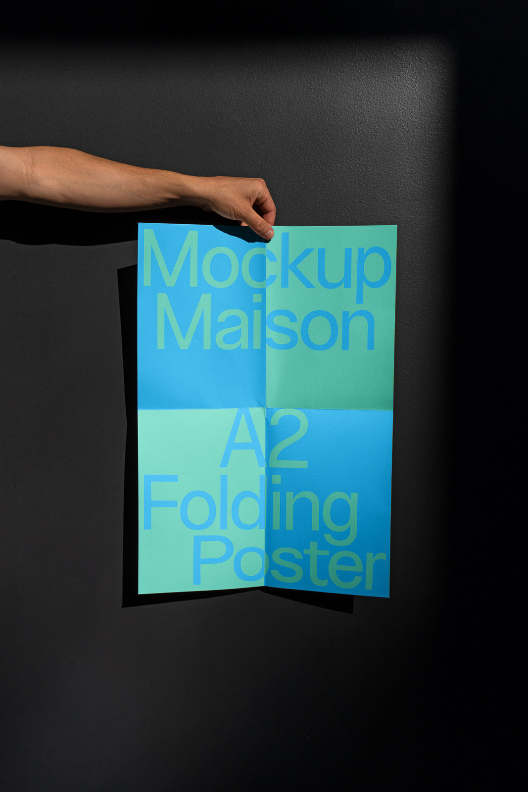PSD mockup of a hand holding an A2 folding poster against a dark background, displaying the text "Mockup Maison" in large blue letters.