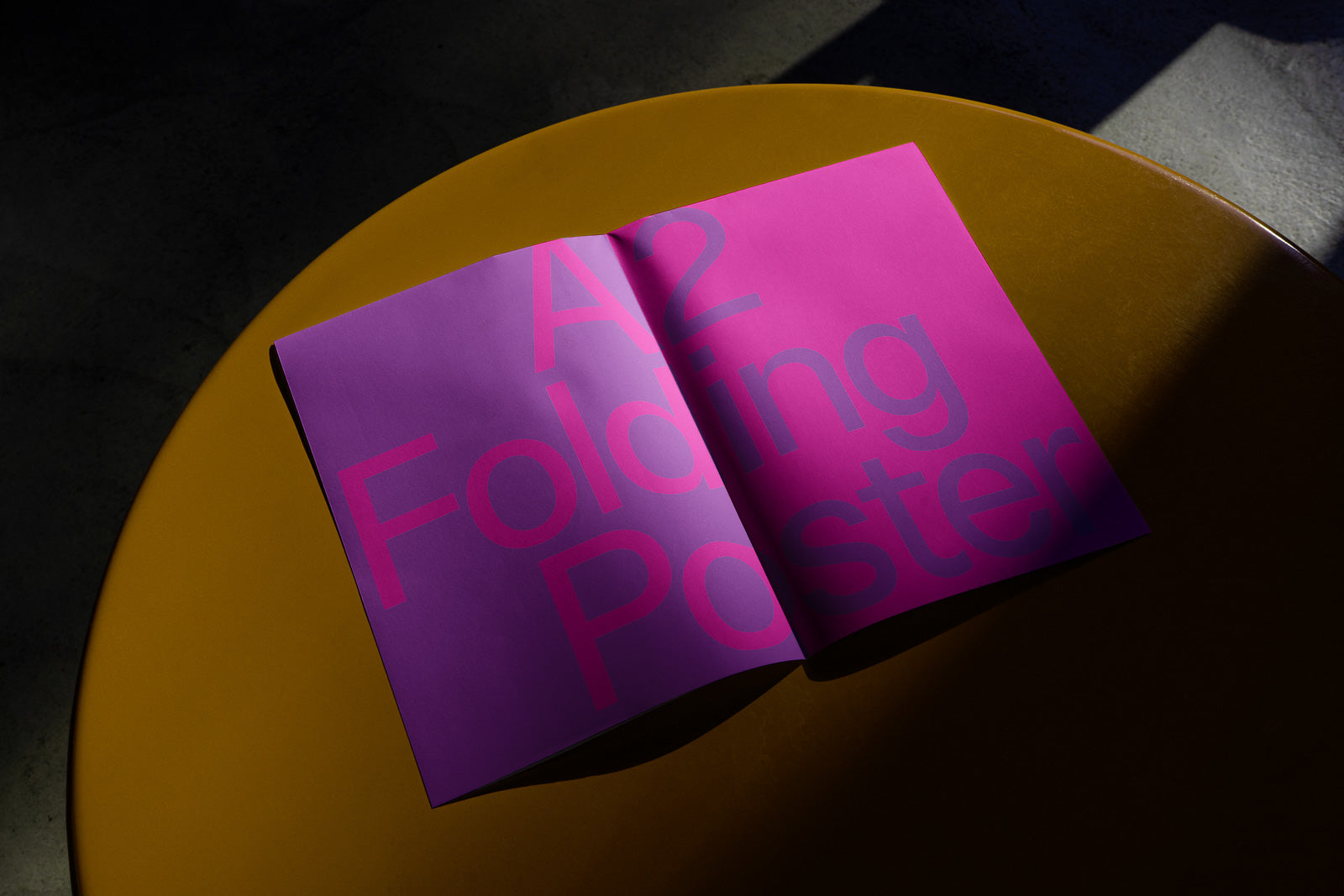 PSD mockup of an open A2 folding poster with bold pink and purple text, set on a circular orange table under dramatic lighting.